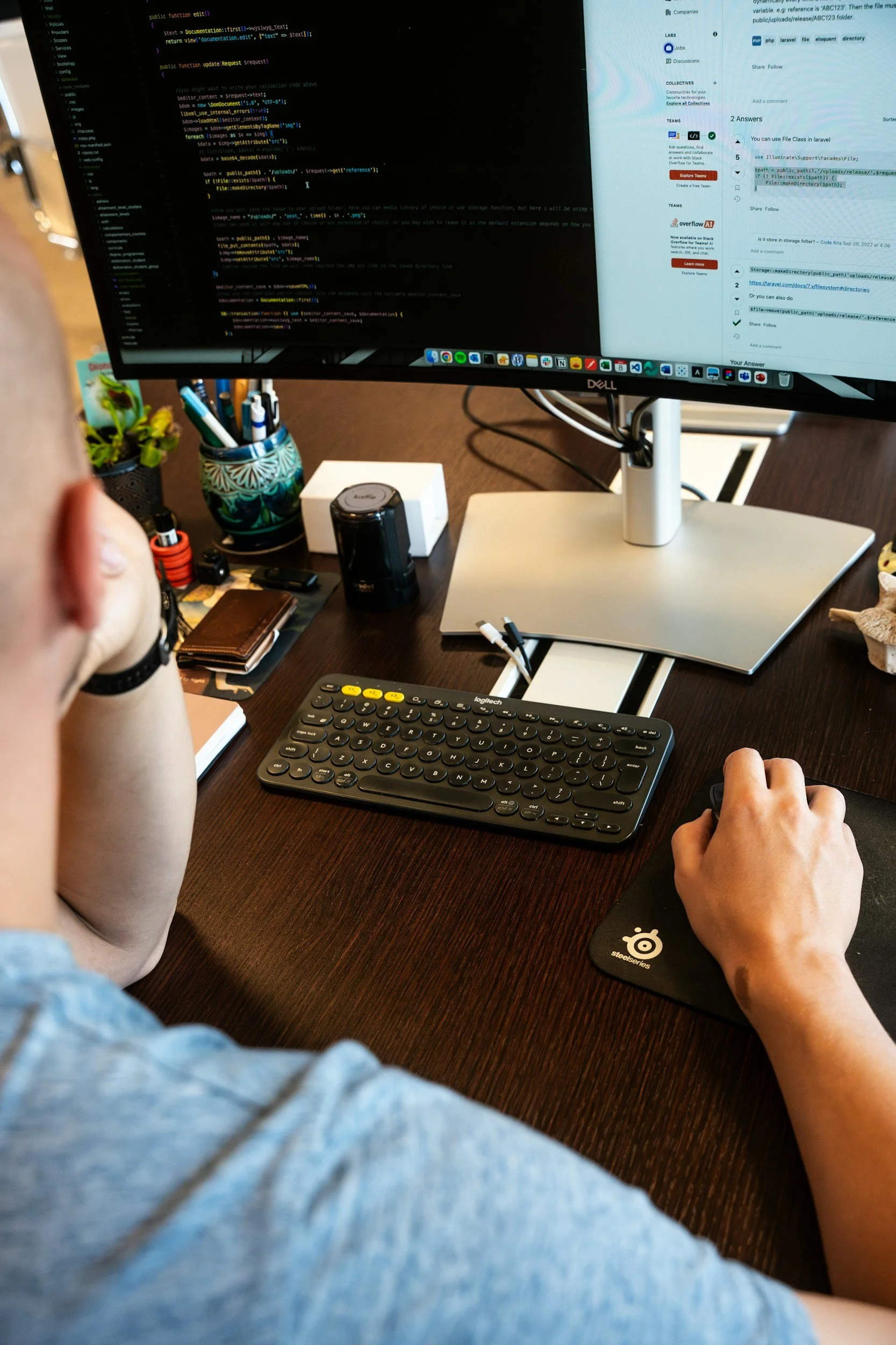 A person working at a desk with a computer monitor displaying coding and web content. The desk has a keyboard, mouse, pen holder, small plant, wallet, and various office supplies.