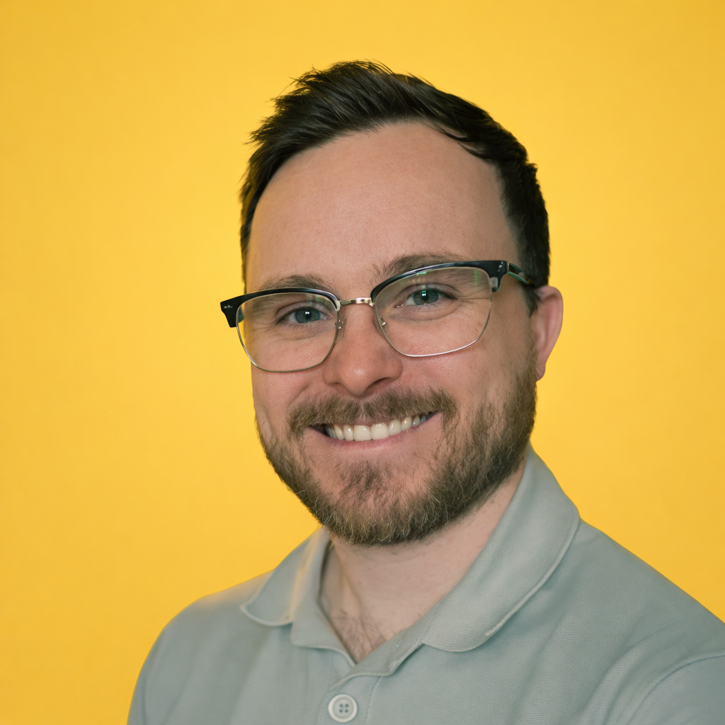 A smiling man with glasses, a beard, and short dark hair, wearing a light gray polo shirt, posing in front of a yellow background.
