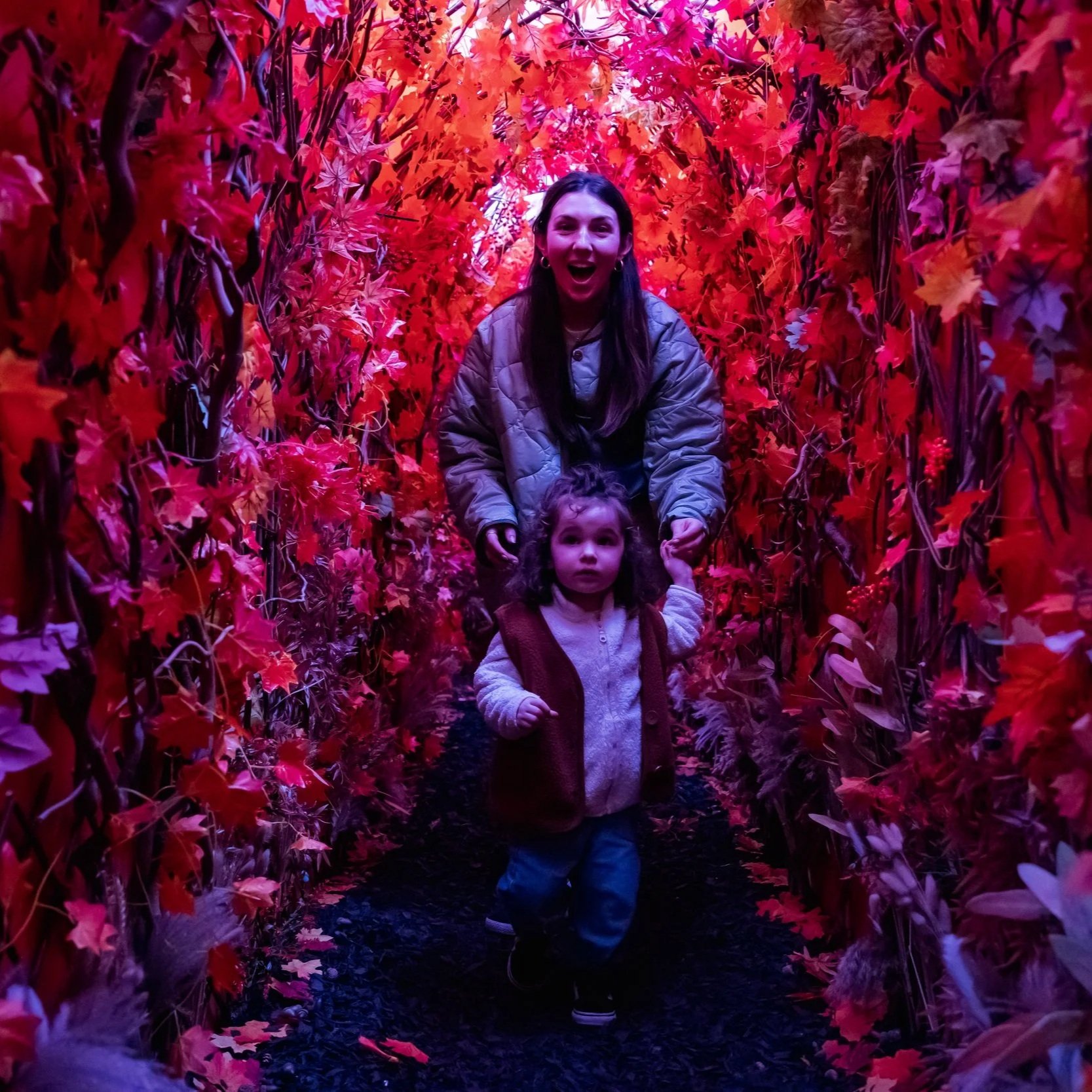 A woman and a young girl walking through a tunnel of red and orange artificial autumn leaves with purple lighting.