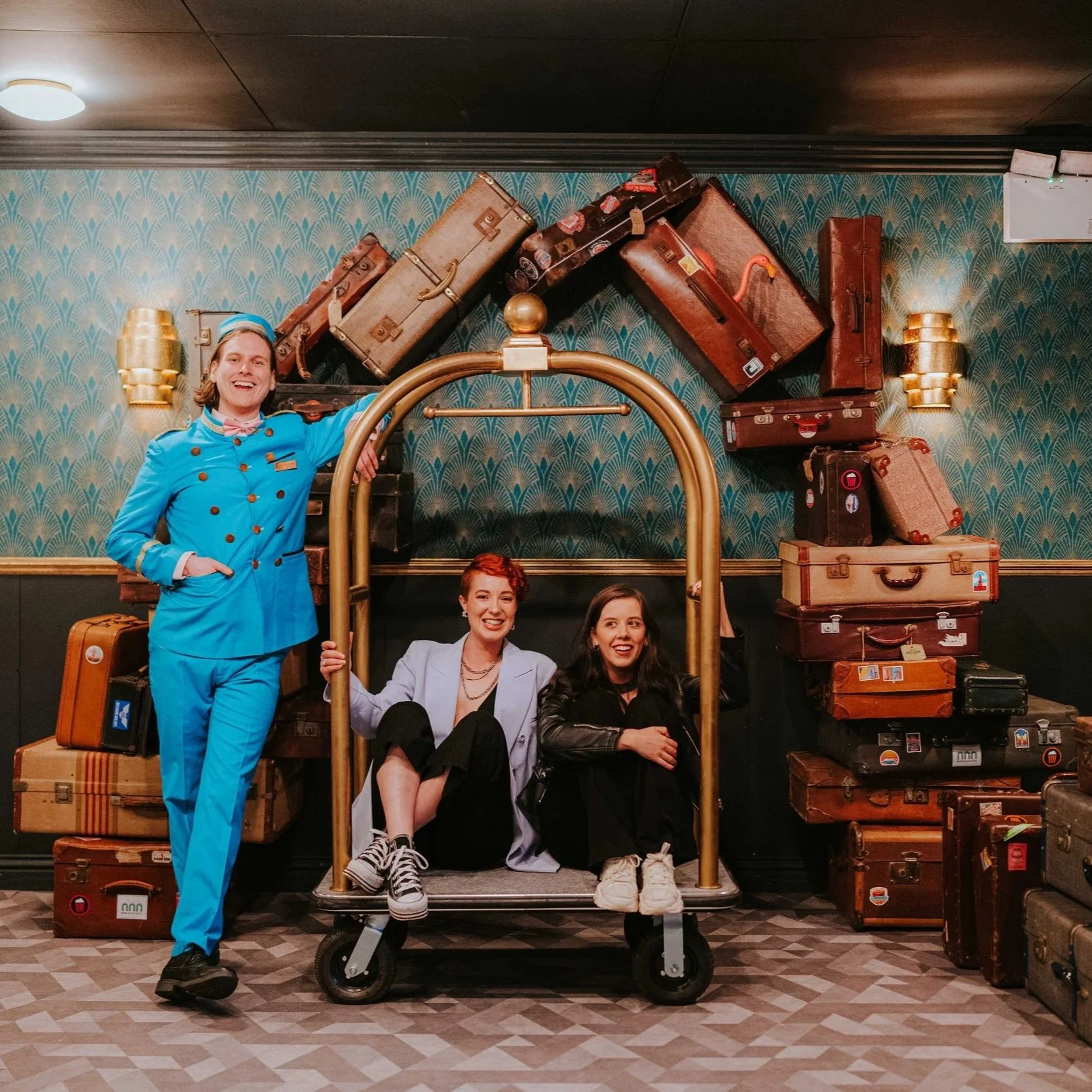 People posing with vintage suitcases in a hotel or luggage store, with an ornate patterned wall and golden wall sconces in the background.