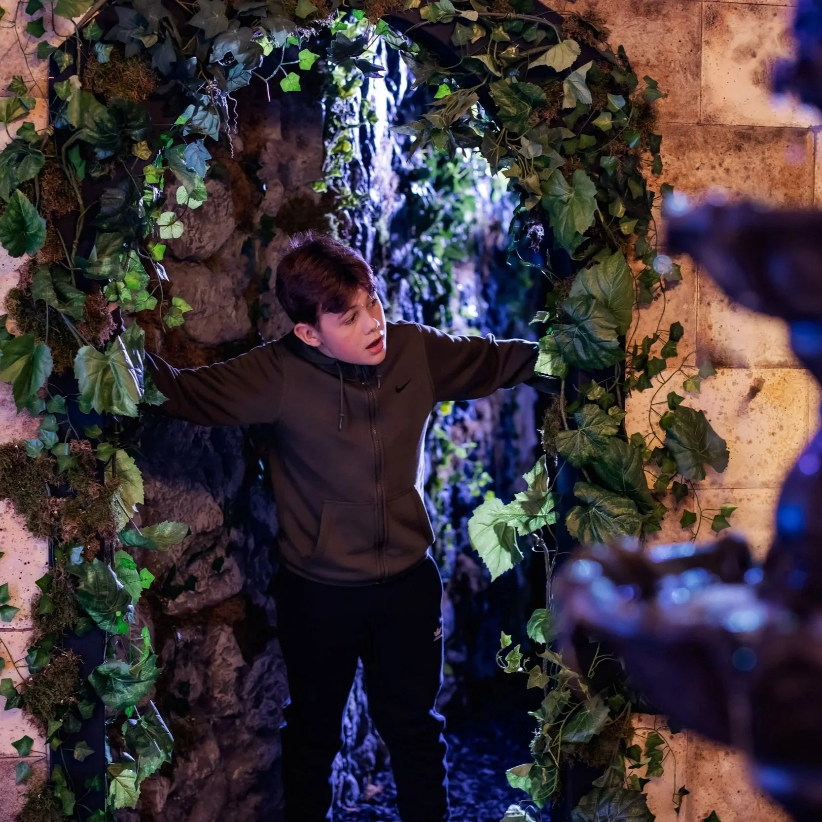 A young boy in a dark brown Nike jacket standing inside a cave-like structure surrounded by green ivy and leaves, with rocky walls and dim lighting.