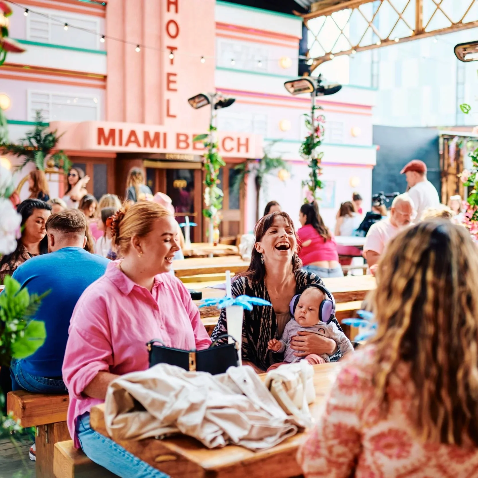 A group of people sitting at picnic tables inside a bright, colorful restaurant or cafe with pink and teal decor, string lights, and green plants. Some are smiling and engaging, including a woman holding a baby wearing headphones.