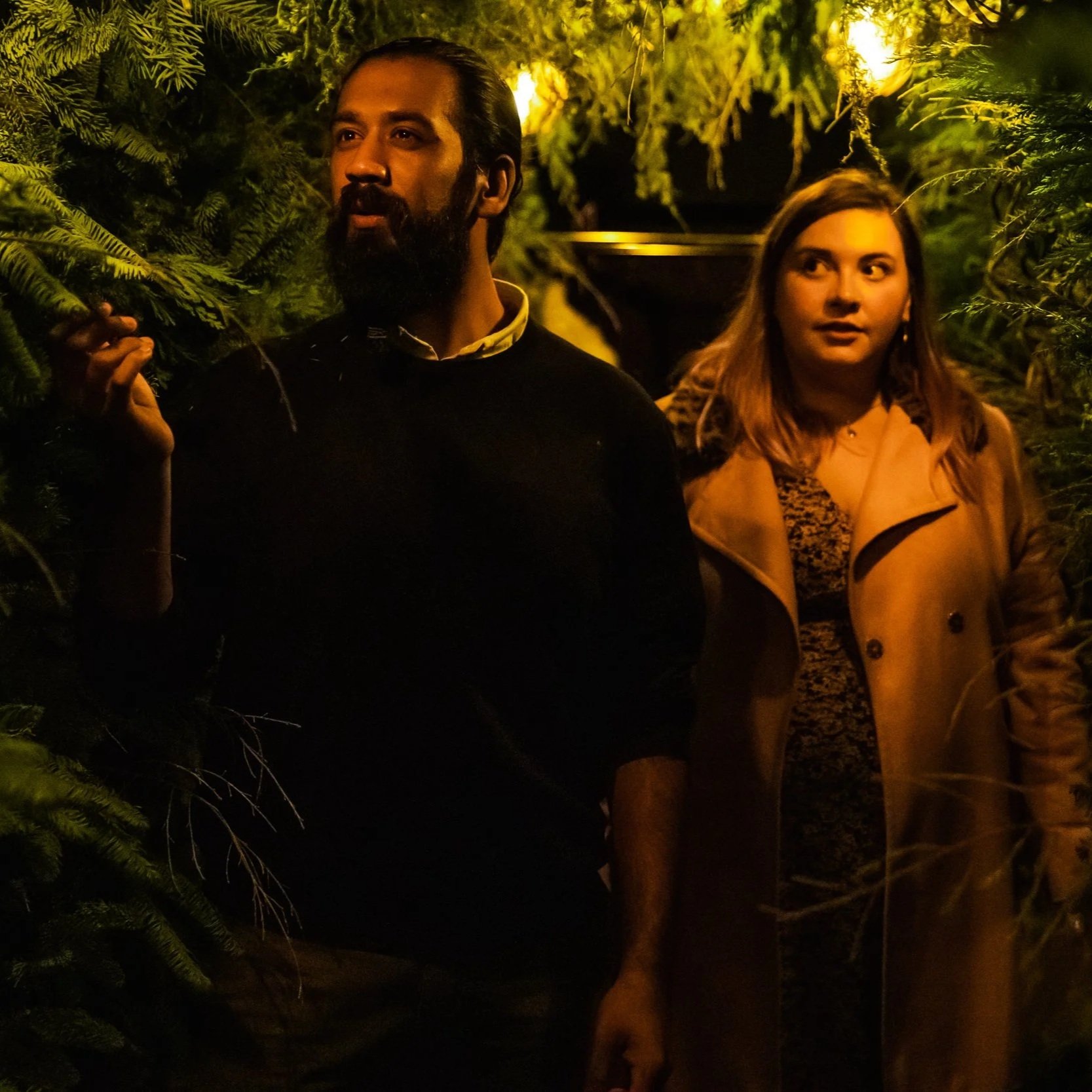 Two people, a man and a woman, standing outdoors at night surrounded by greenery and illuminated by warm yellow lighting.