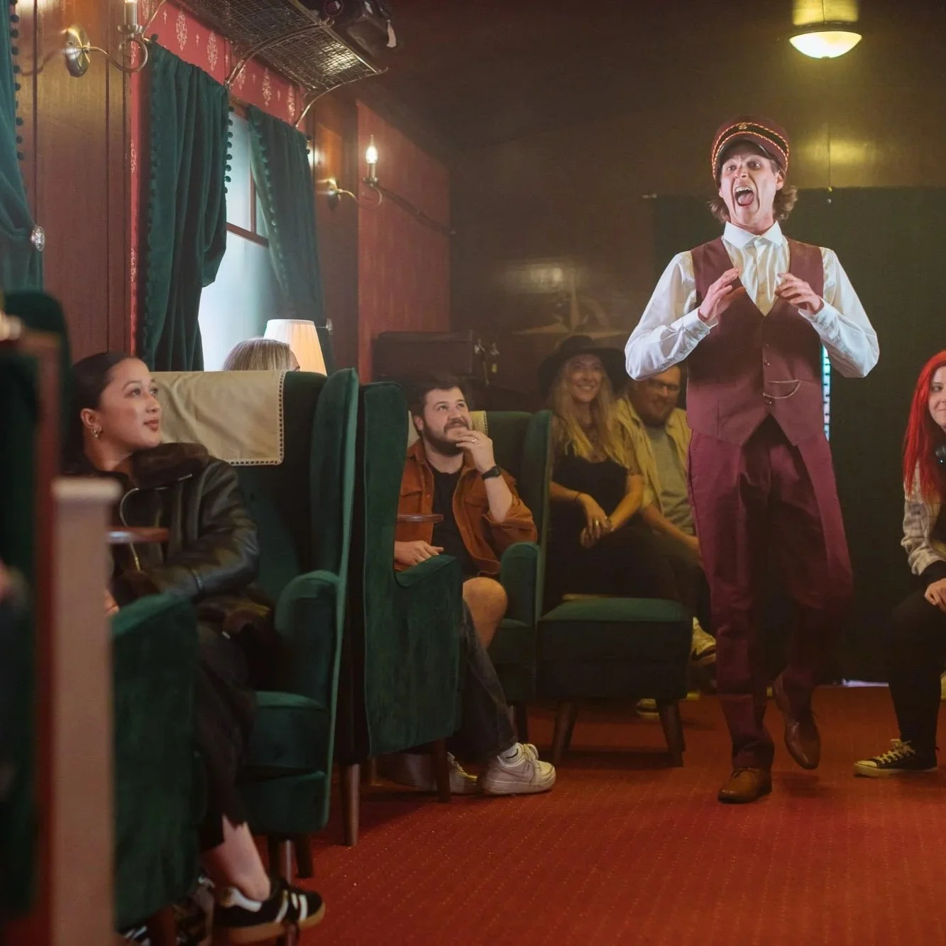 A group of people seated in a cozy, dimly-lit room with green velvet chairs and dark wooden walls, watching a performer with a mustache and brown vest who appears to be acting or speaking animatedly.