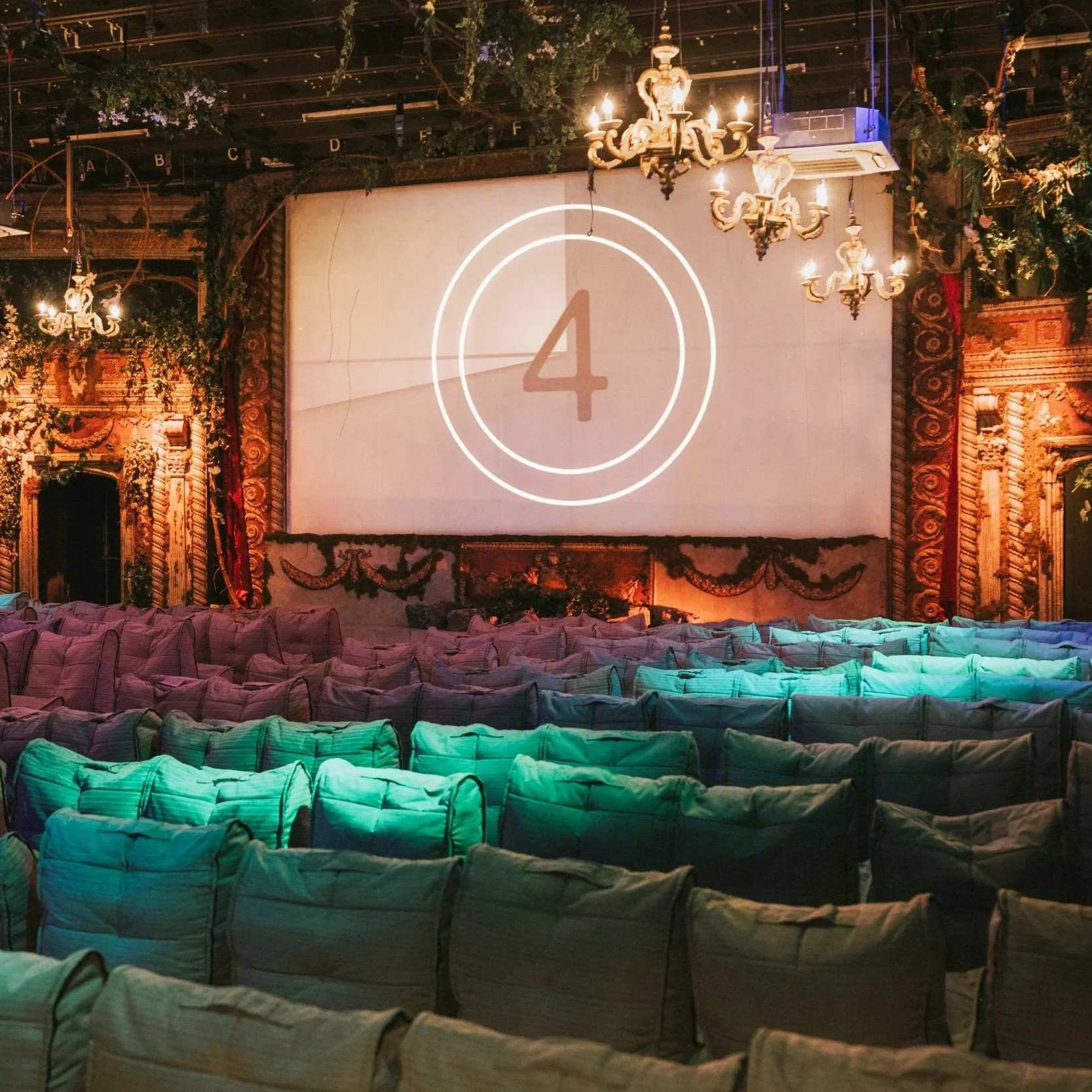 An empty stage with a large screen displaying a countdown timer at four seconds, decorated with chandeliers, floral arrangements, and ornate gold detailing, and rows of chairs covered in fabric in front.