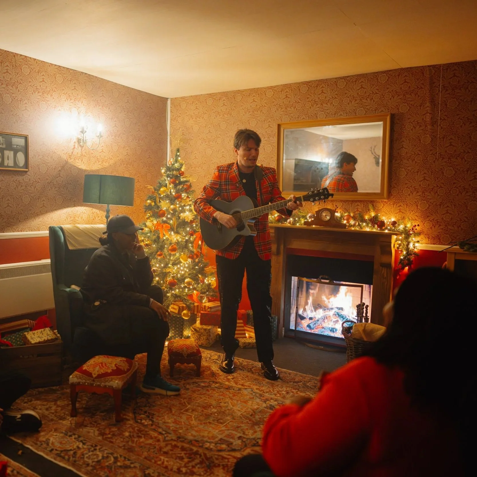 A man in a red plaid jacket playing the guitar in a cozy Christmas living room, decorated with a Christmas tree, garlands, and a lit fireplace.