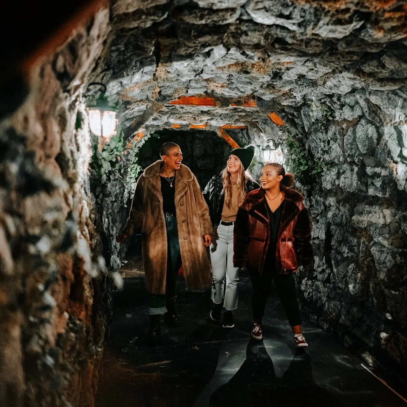 Three women walking and talking inside a tunnel with stone walls, smiling and wearing fall clothing.