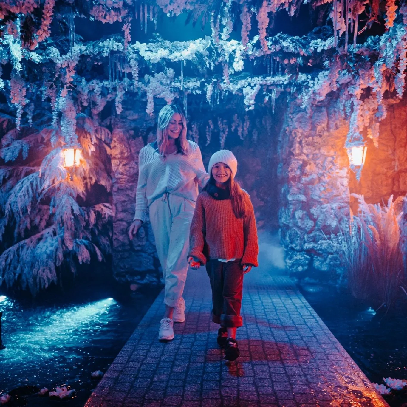 A woman and a young girl walk on a illuminated pathway in an indoor winter-themed setting with snow-covered plants and rocks, and warm orange and blue lighting.