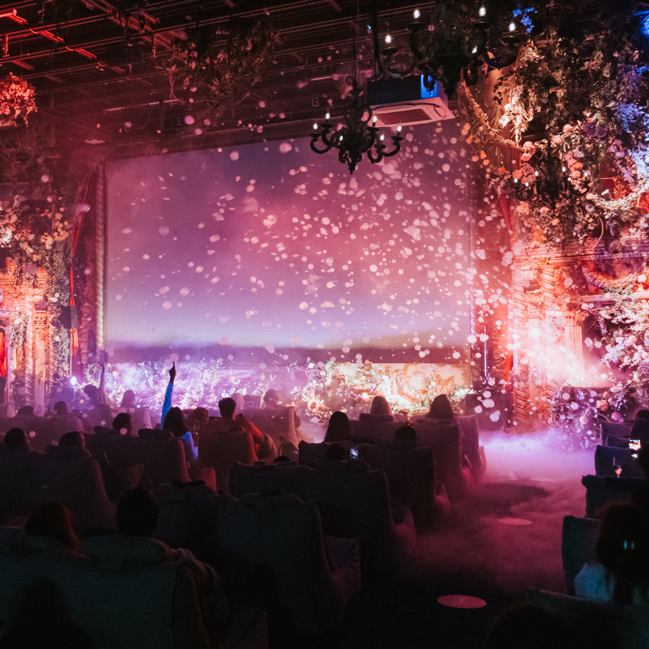People sitting in an auditorium watching a colorful light show on a large screen, with fog and decorative lighting.