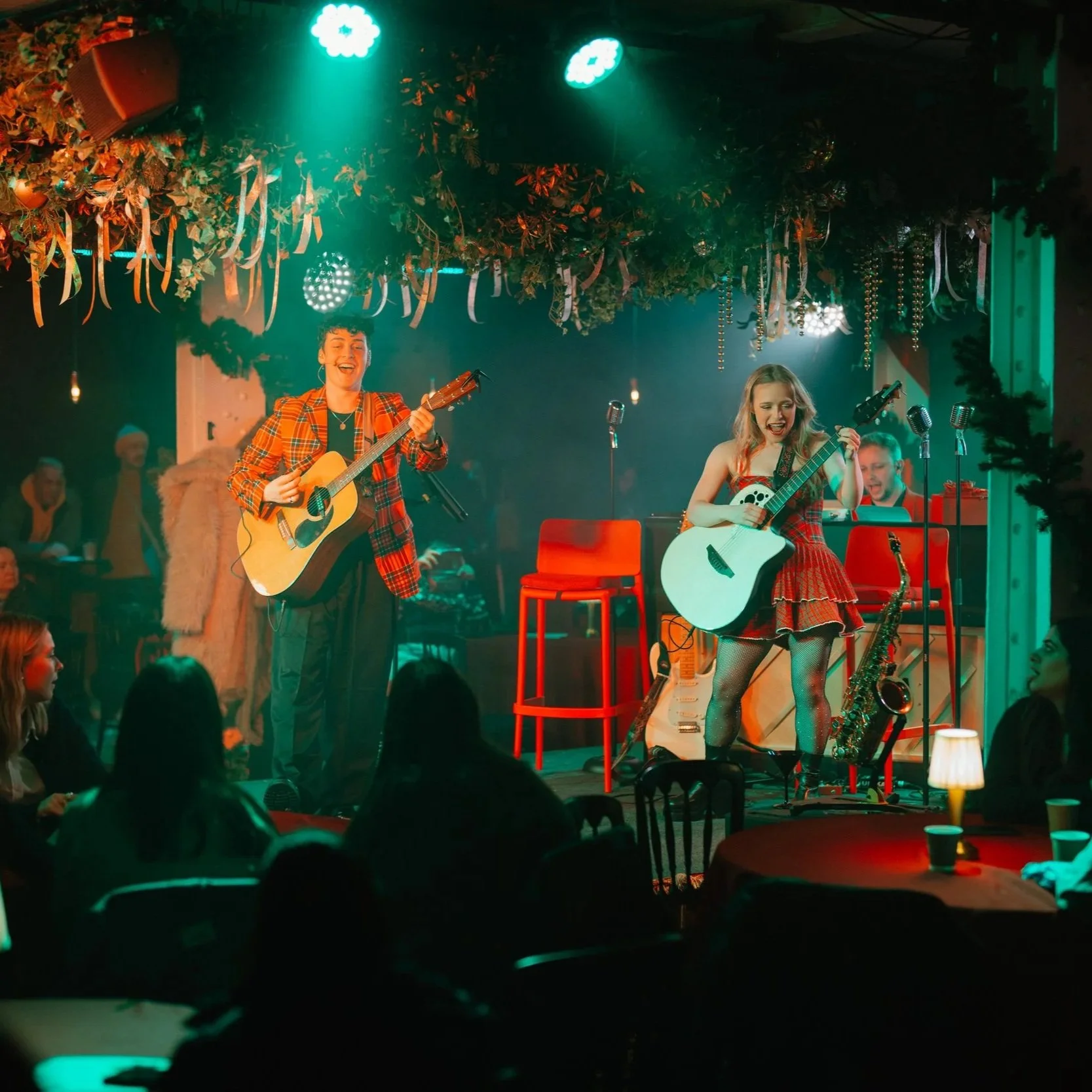 Two women on stage playing guitars, one singing, surrounded by Christmas decorations, audience in foreground, stage lit with green lights.