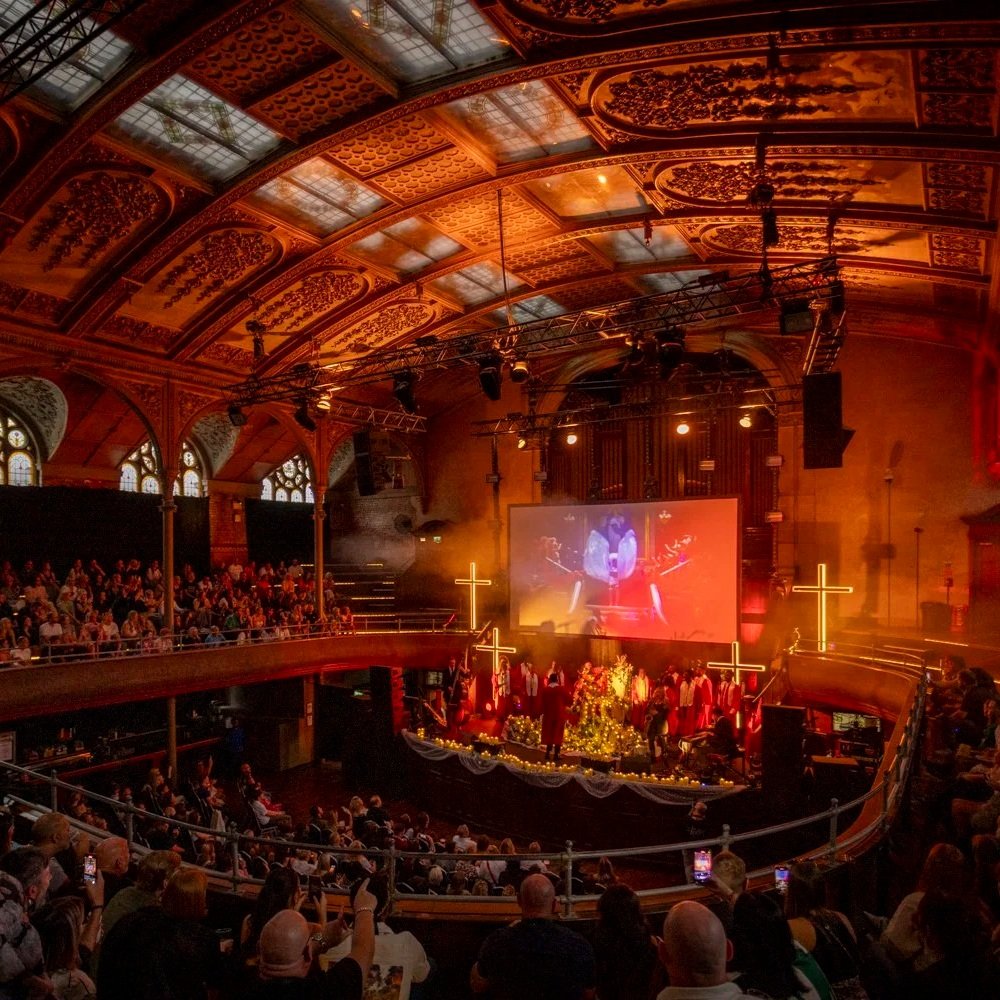 Inside a church with a high, decorated ceiling and stained glass windows, a large congregation is attending a Christmas or holiday event featuring a choir on stage, Christmas decorations, and illuminated crosses.