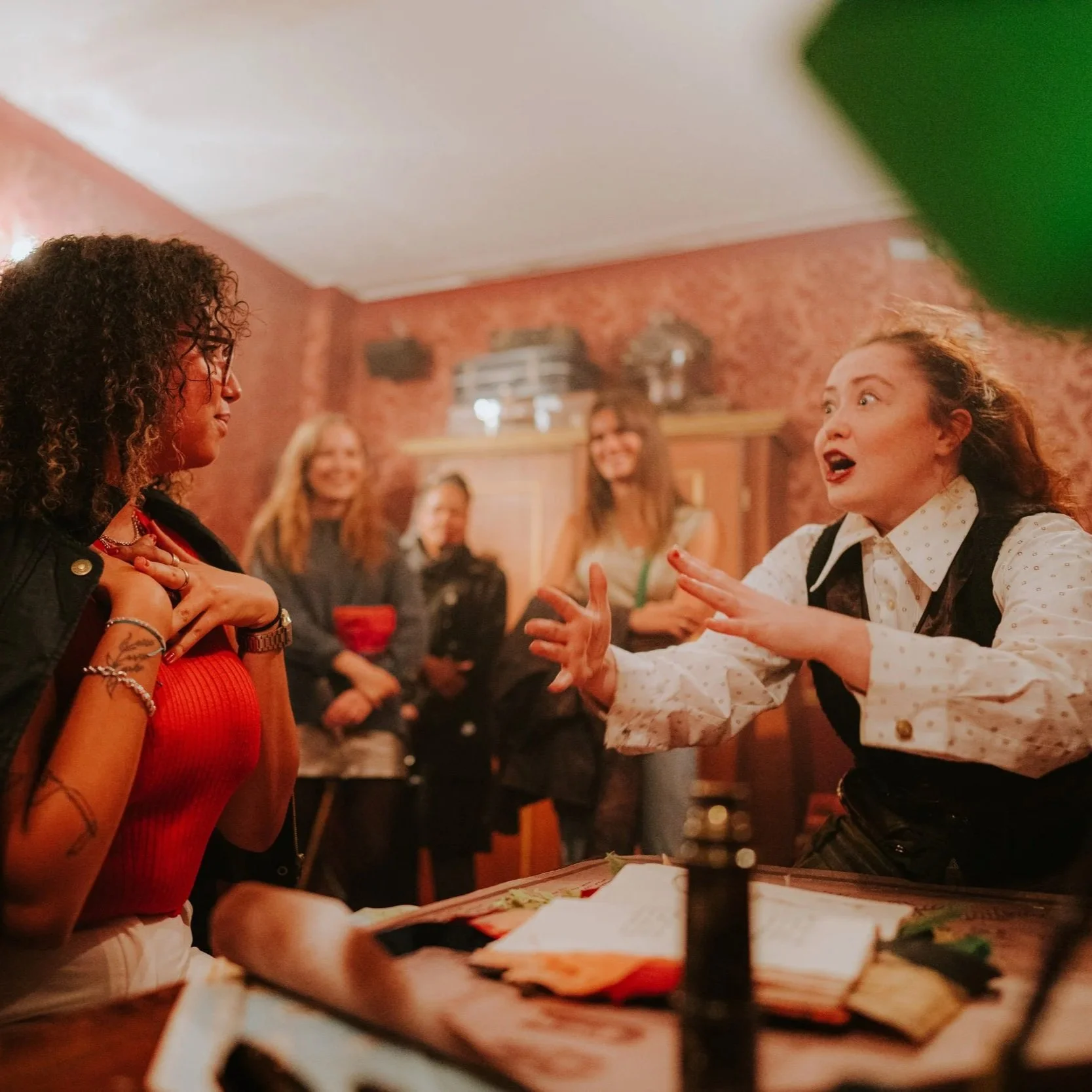 A group of women socializing in a cozy room with warm lighting and patterned wallpaper. Two women in the foreground are engaged in conversation, one with curly hair and the other with reddish hair tied back, expressing animated emotion. Several other