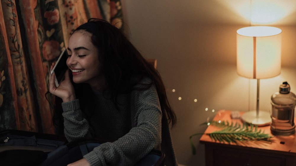 A young woman with long dark hair, smiling, talking on a phone while sitting at a desk in a warmly lit room with curtains and a bedside table with a lamp, glass jar, and green leaf plants.