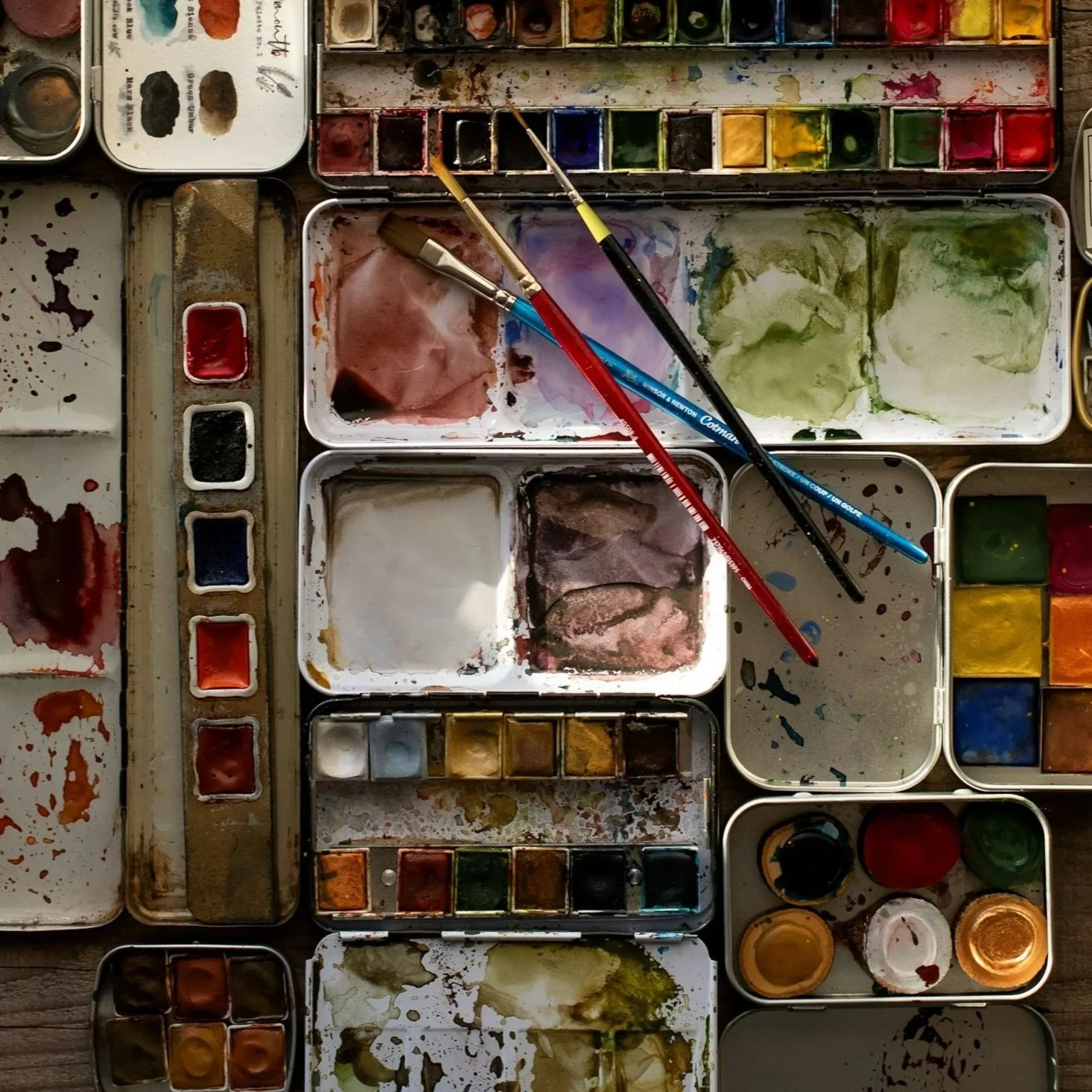 A collection of watercolor paint palettes, brushes, and mixed colors on a wooden surface.