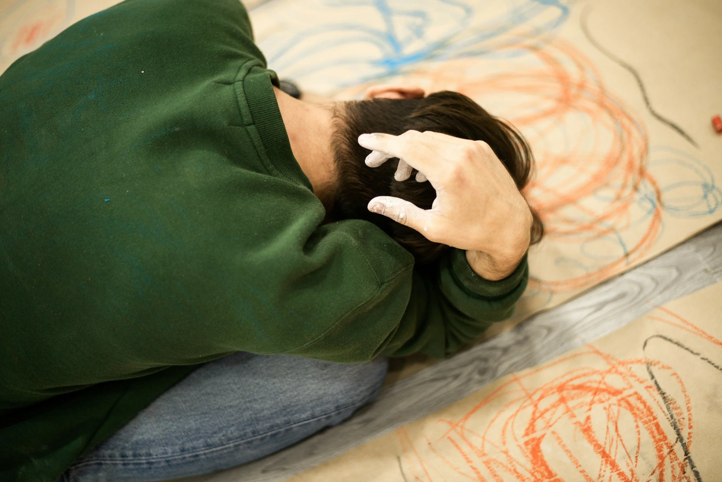 Person lying on the floor with head resting on their knees, face covered by hands, in front of colorful chalk drawings on large sheets of paper.