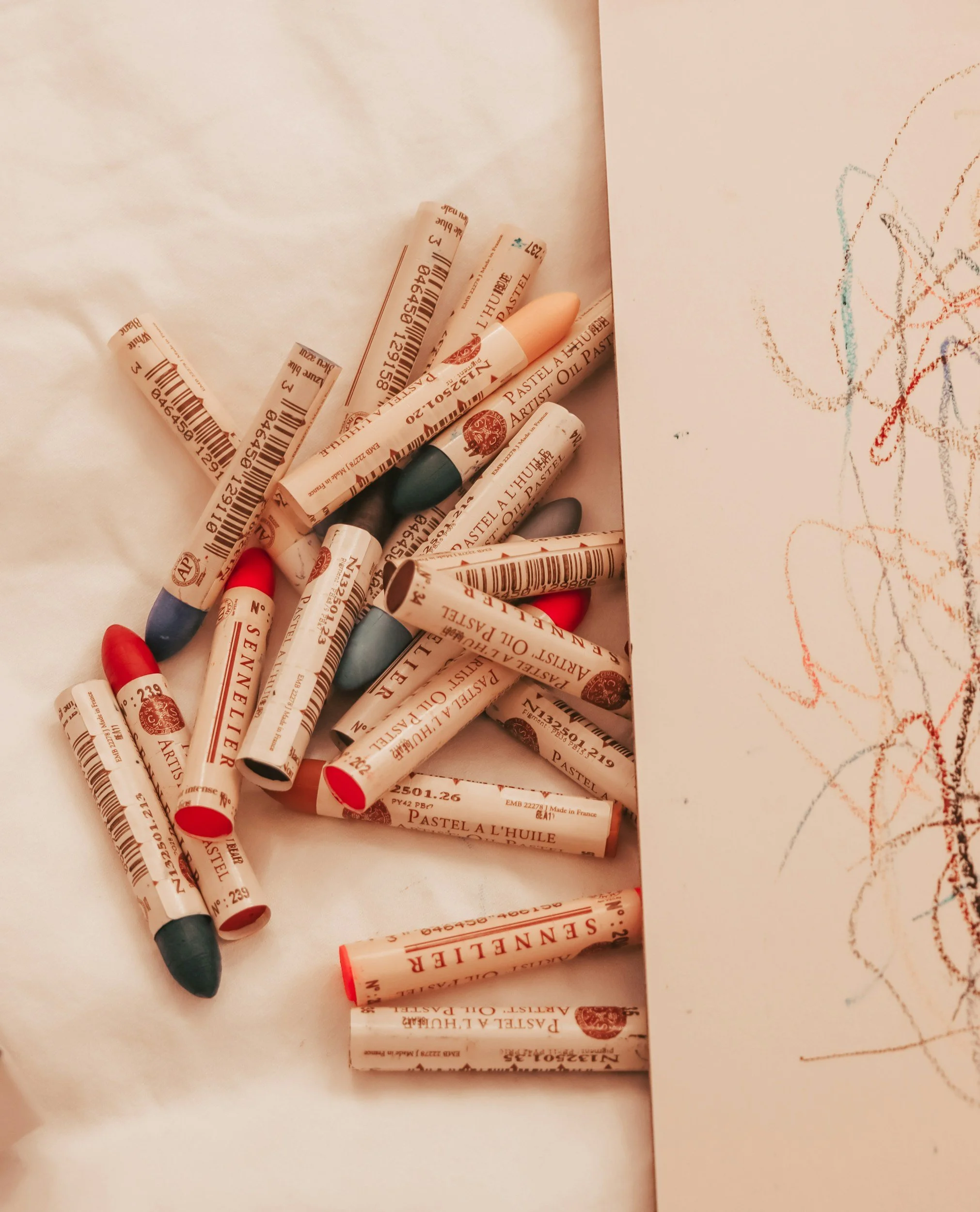 Assorted oil pastels, some with paper labels, scattered on white paper, with a child's crayon drawing on the right.
