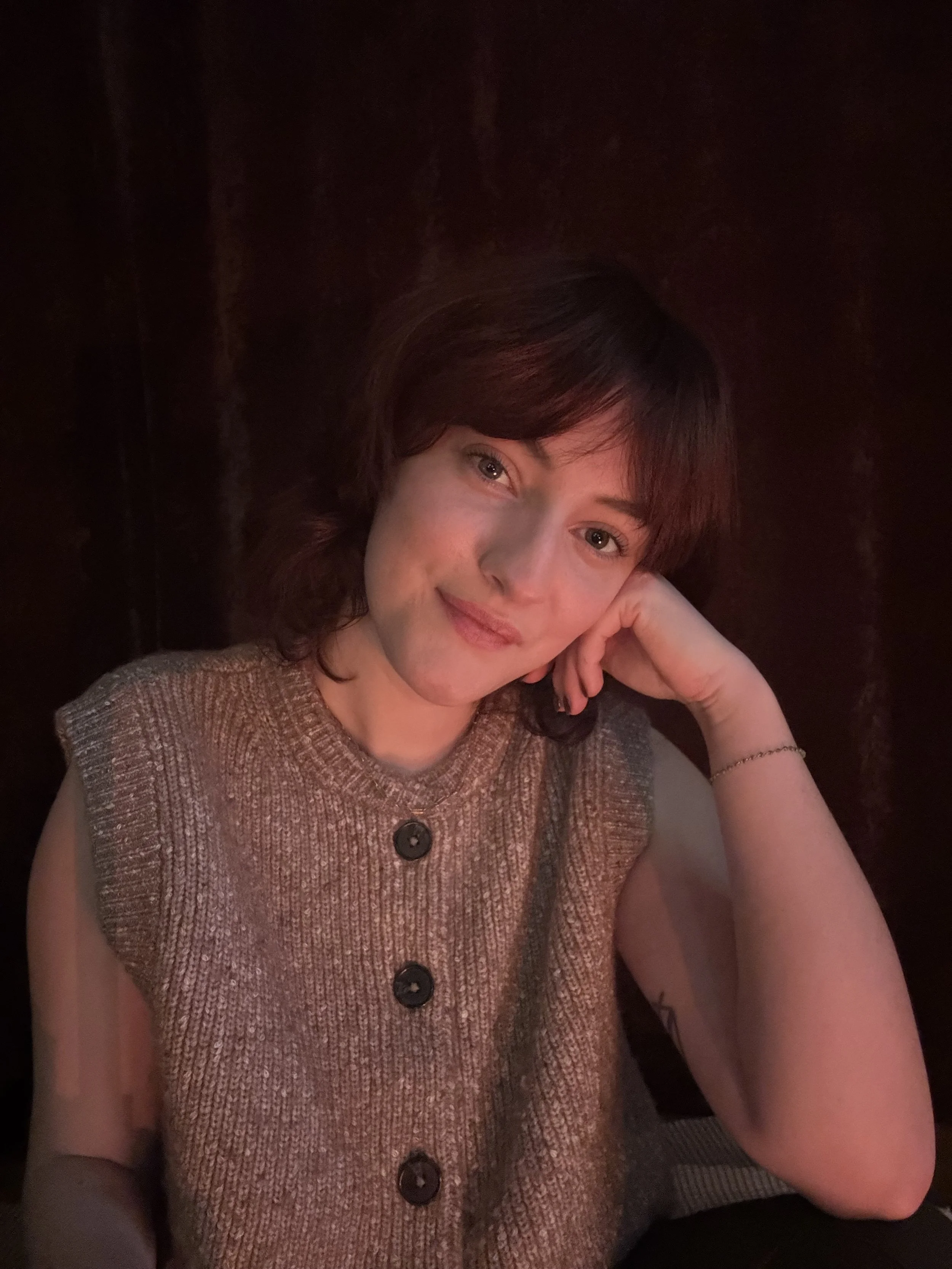 A woman with short brown hair and light skin, resting her head on her hand, smiling softly at the camera. She is wearing a sleeveless, knitted brown vest with large buttons, and a thin bracelet on her wrist. The background is dark and blurred.
