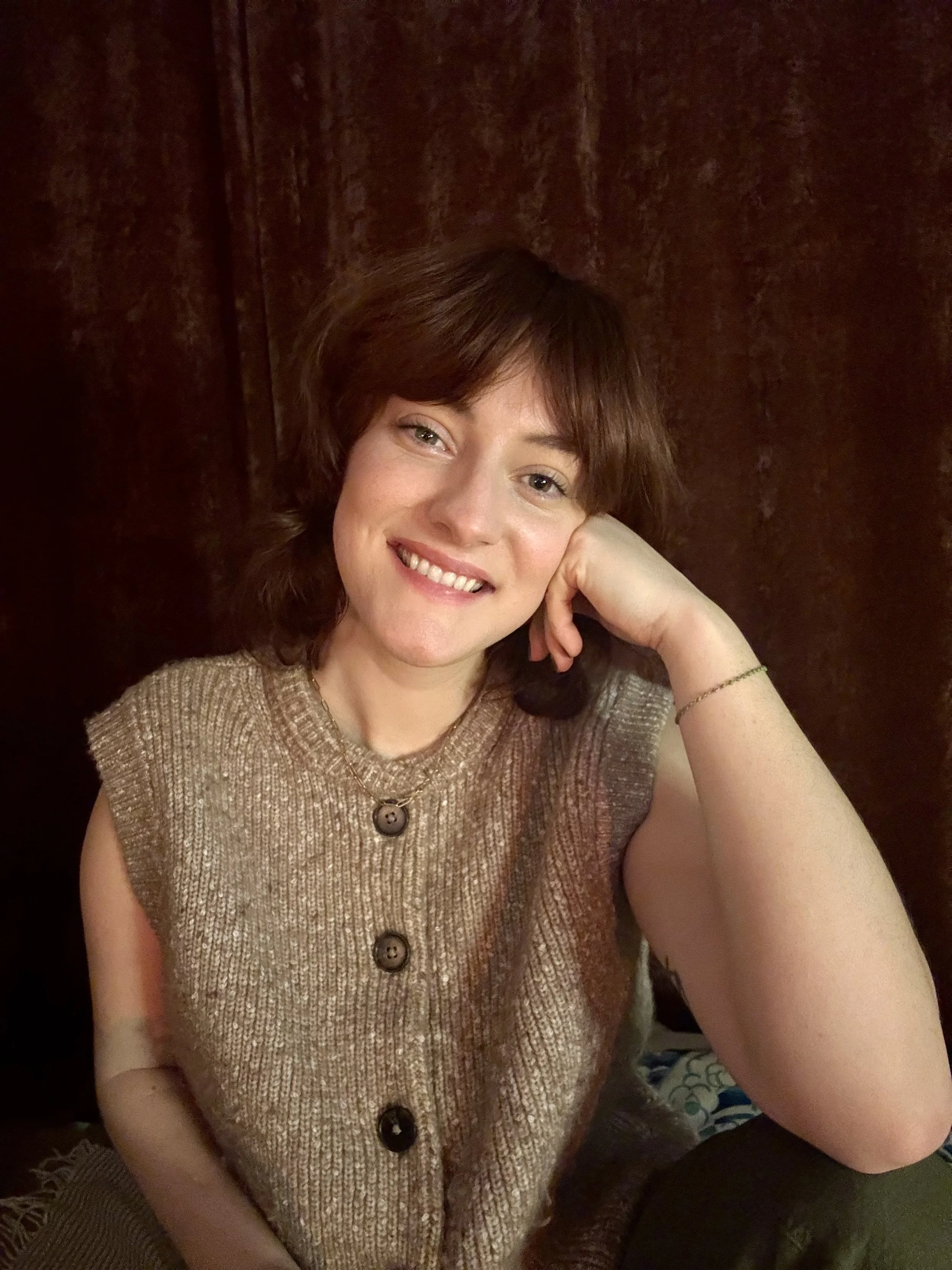 A woman with short brown hair and fair skin smiling, resting her head on her hand, wearing a sleeveless, knit beige top with large buttons.