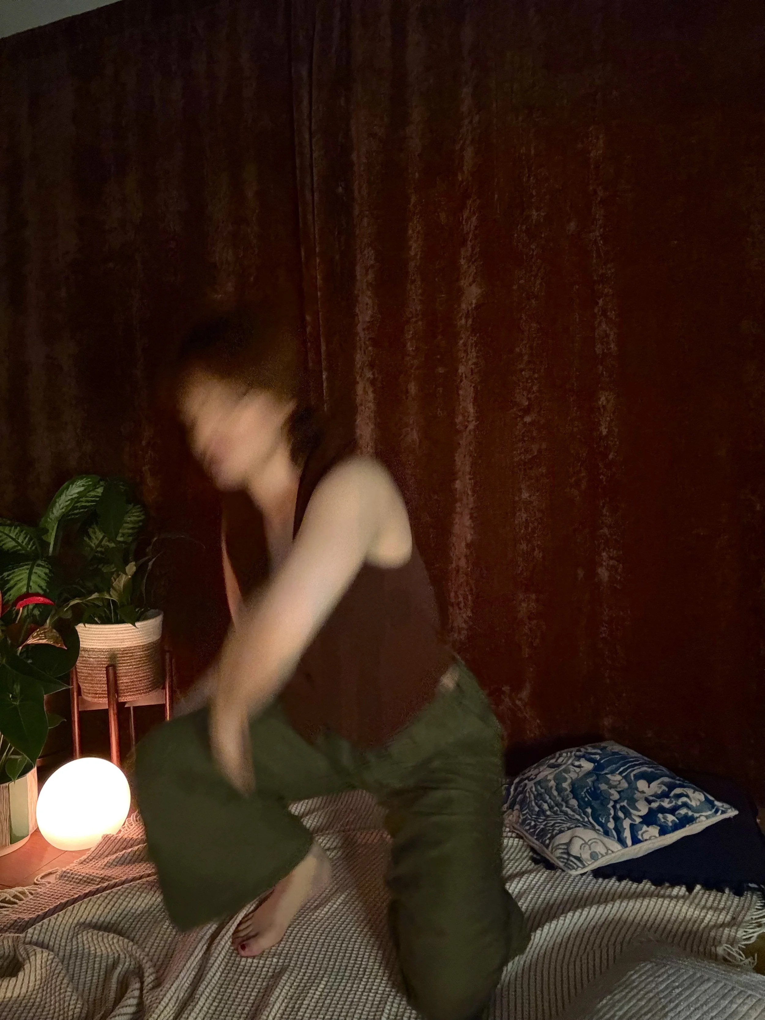 A person with short hair dancing or jumping on a bed in a dimly lit room with red curtains, a potted plant, a glowing lamp, and a pillow with a blue and white design.