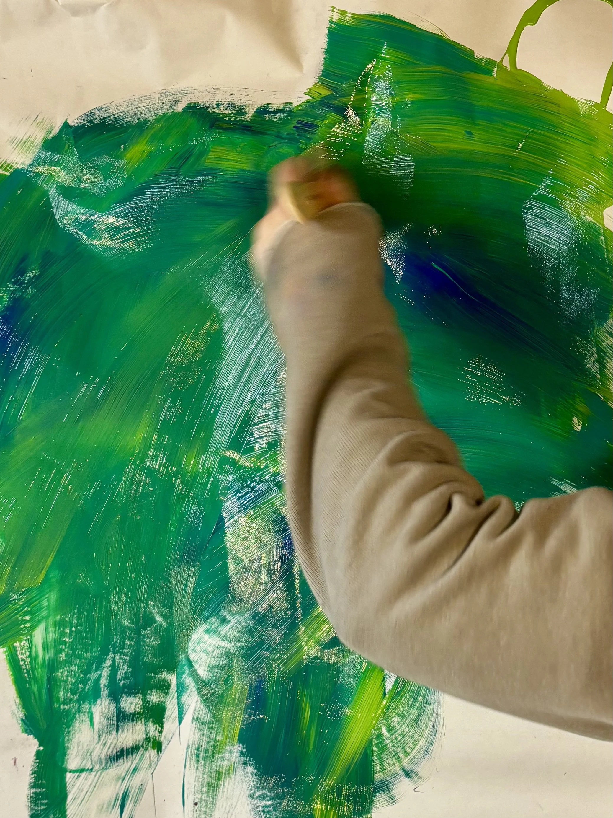 A person creating abstract art with green and blue paint on paper, using a large brush.