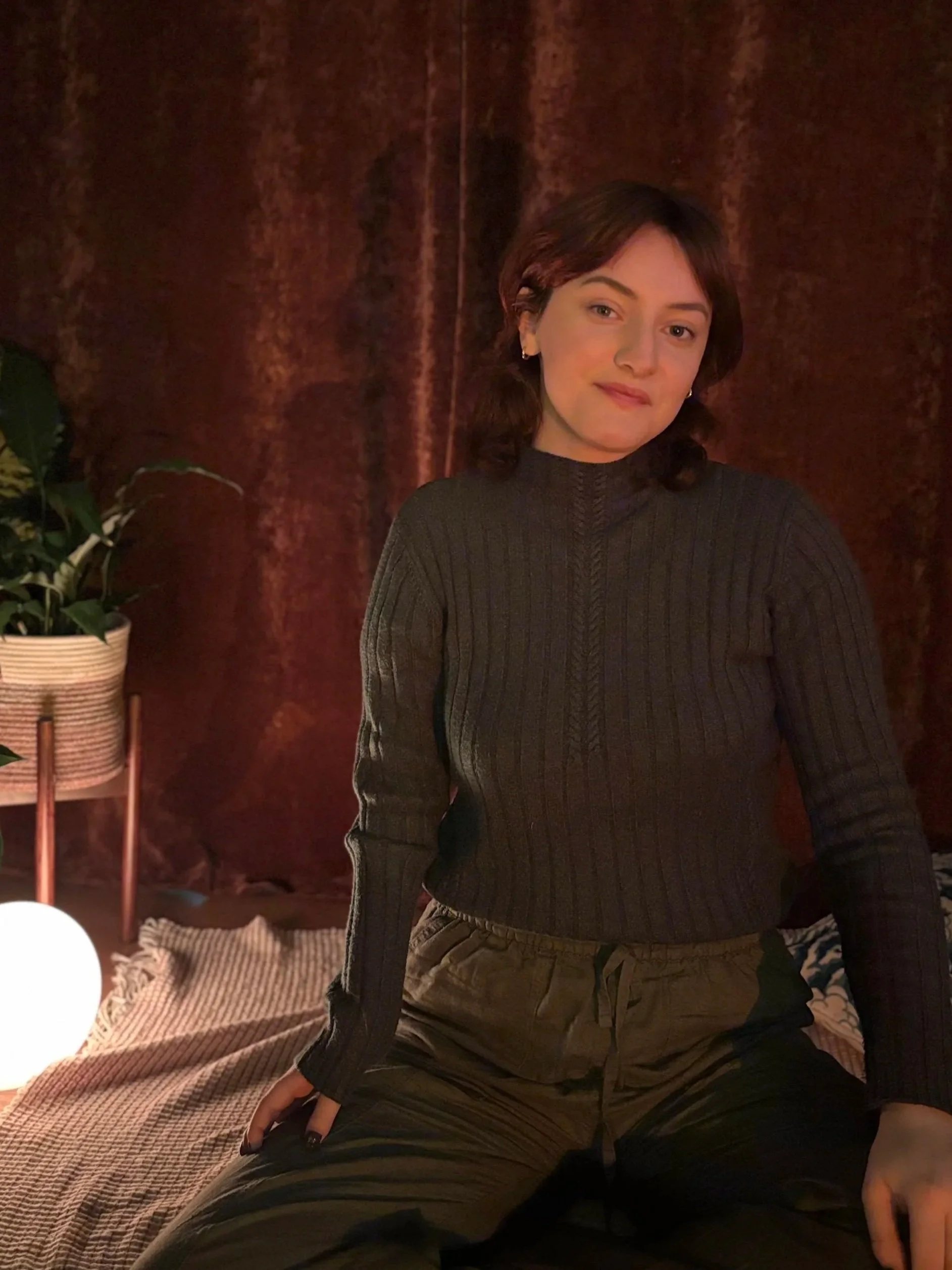 A woman with short brown hair sitting on a bed with a checkered blanket, in a room with dark brown curtains, a potted plant, and a glowing lamp.