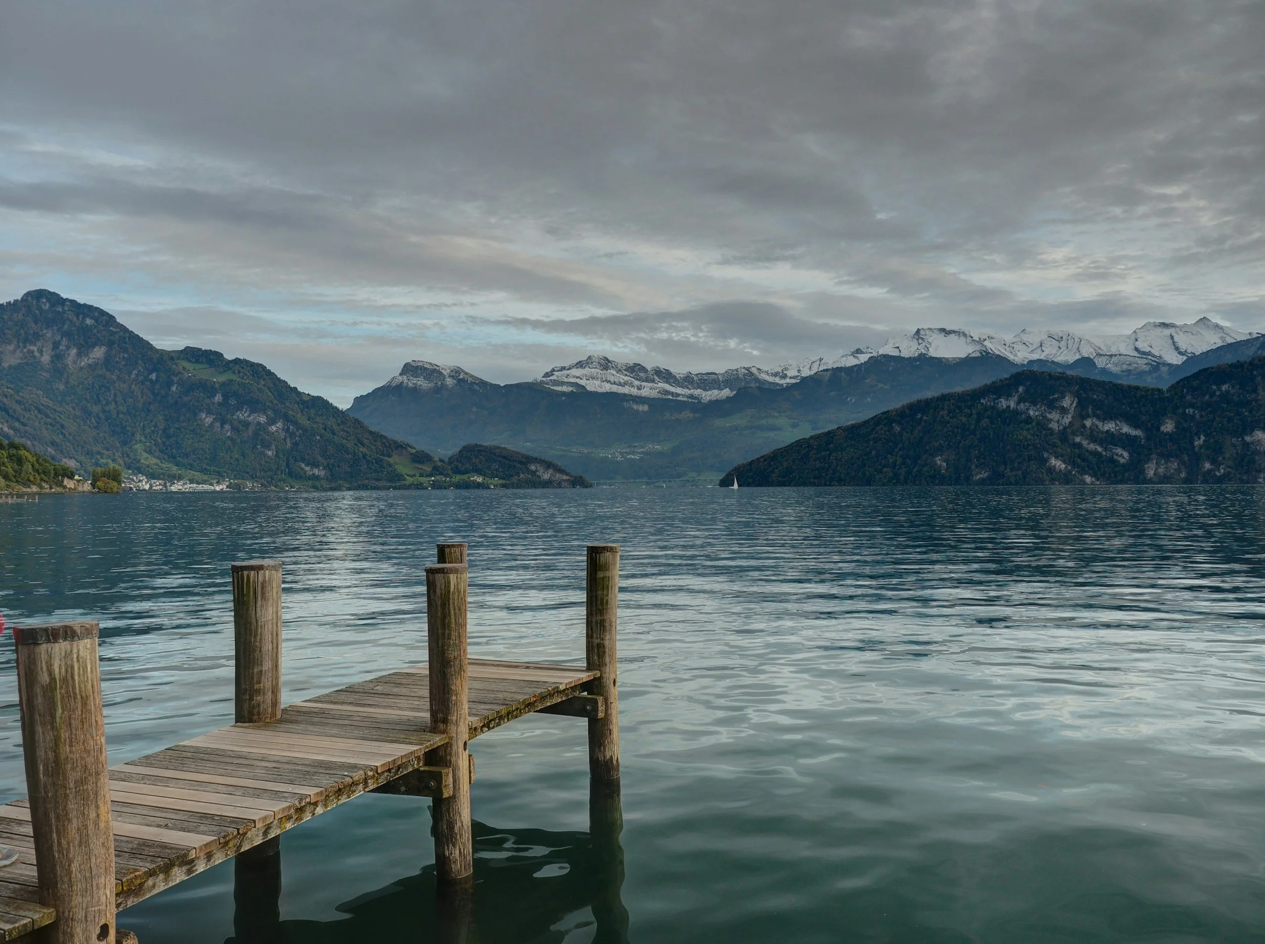 A wooden dock extending into a calm lake surrounded by green hills and snow-capped mountains in the distance under a cloudy sky.