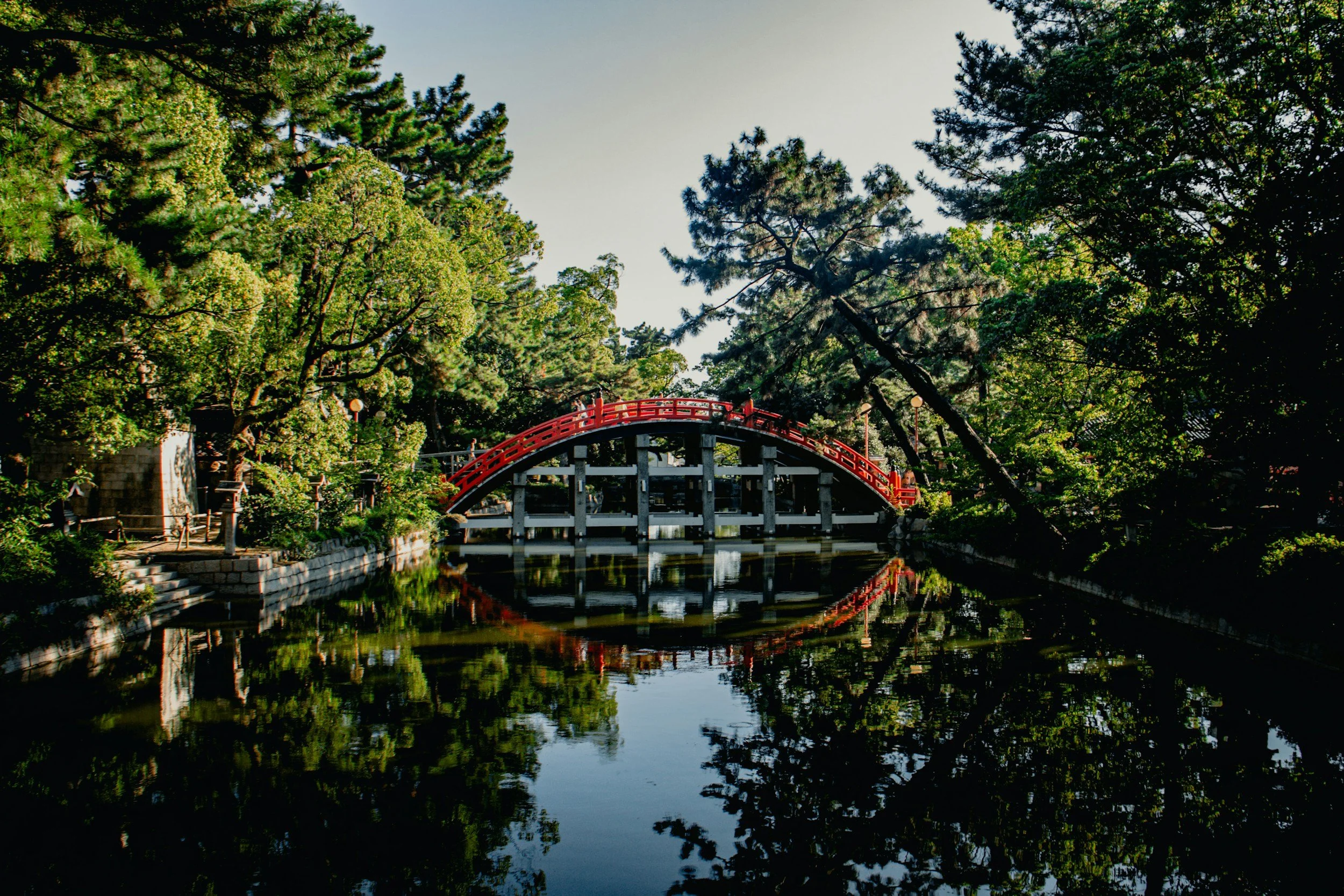 A peaceful scene of a red arched bridge over a calm body of water, surrounded by lush green trees on both sides.