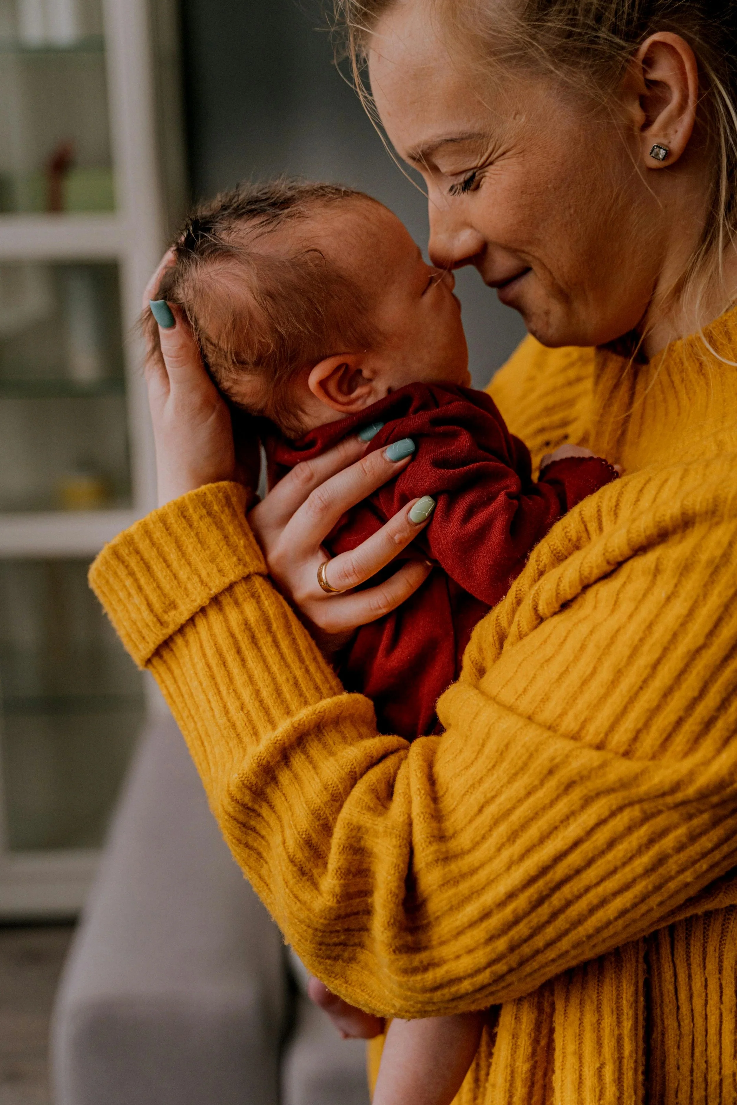 A woman in a mustard yellow sweater lovingly holds a newborn baby close to her face, both with their eyes closed, sharing a tender moment indoors.