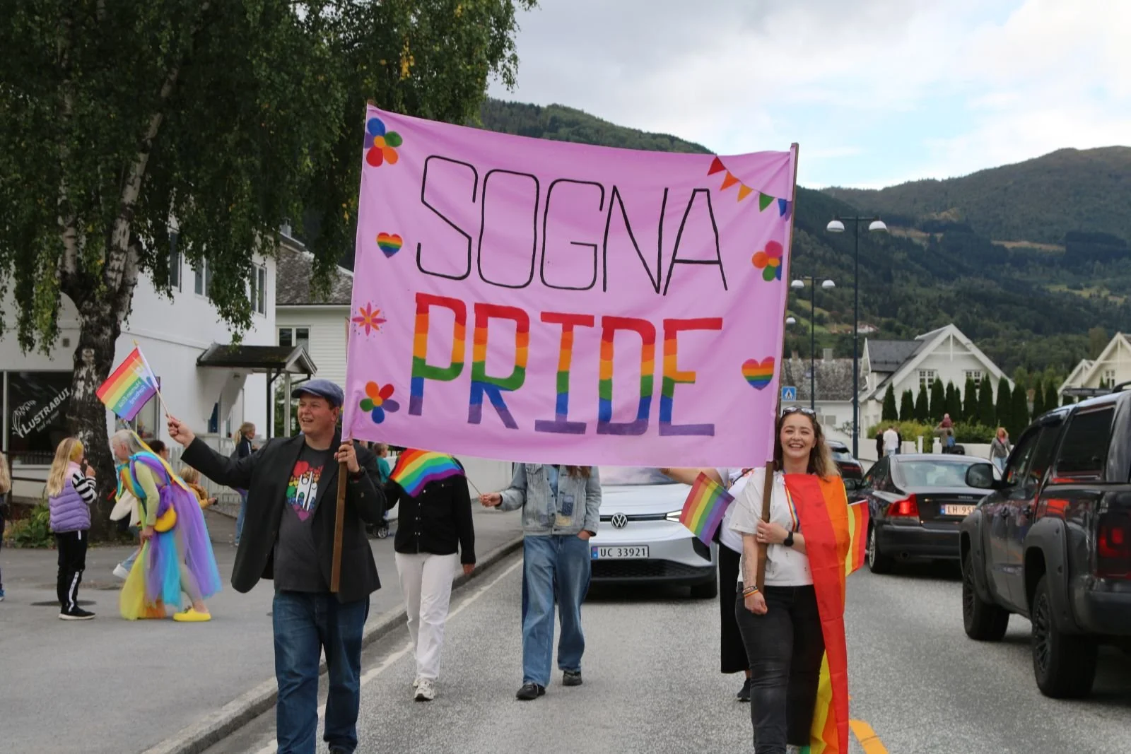 To personer bærer en stor rosa plakat med teksten 'SOGNA PRIDE' og små hjerter og blomster i regnbuefarger, under en parade med andre mennesker og regnbueflagg i en småby med hytter og fjell i bakgrunnen.