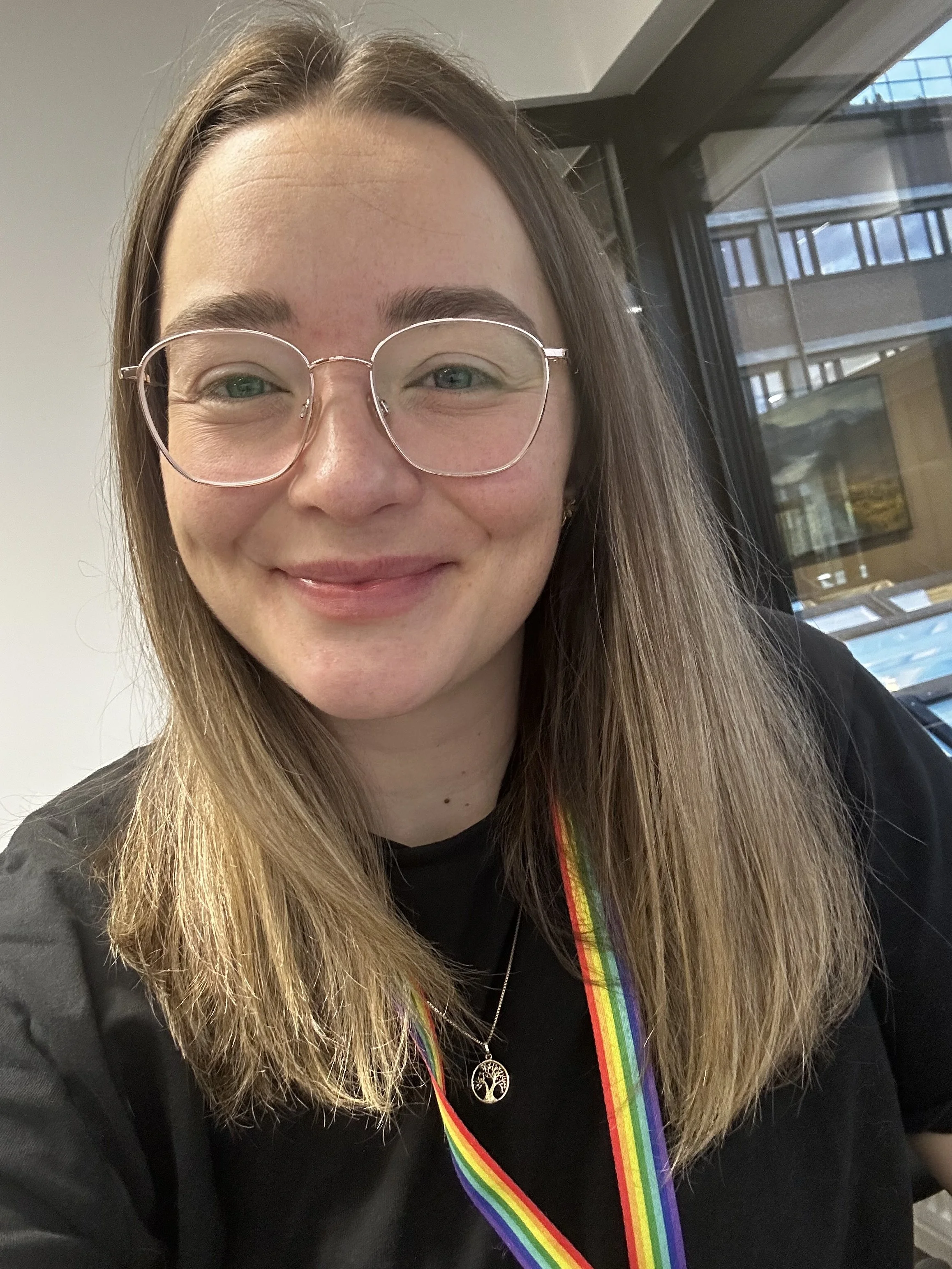 Et smilende bilde av en kvinne med langt, blond hår, briller og en regnbuefarget medaljerekke, sittende inne med et vindu i bakgrunnen.