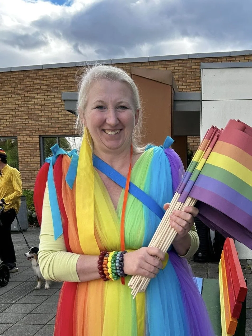 En smilende kvinne kledd i en regnbuefarget kjole, holder små regnbuefargede flagg, på en utendørsområde.
