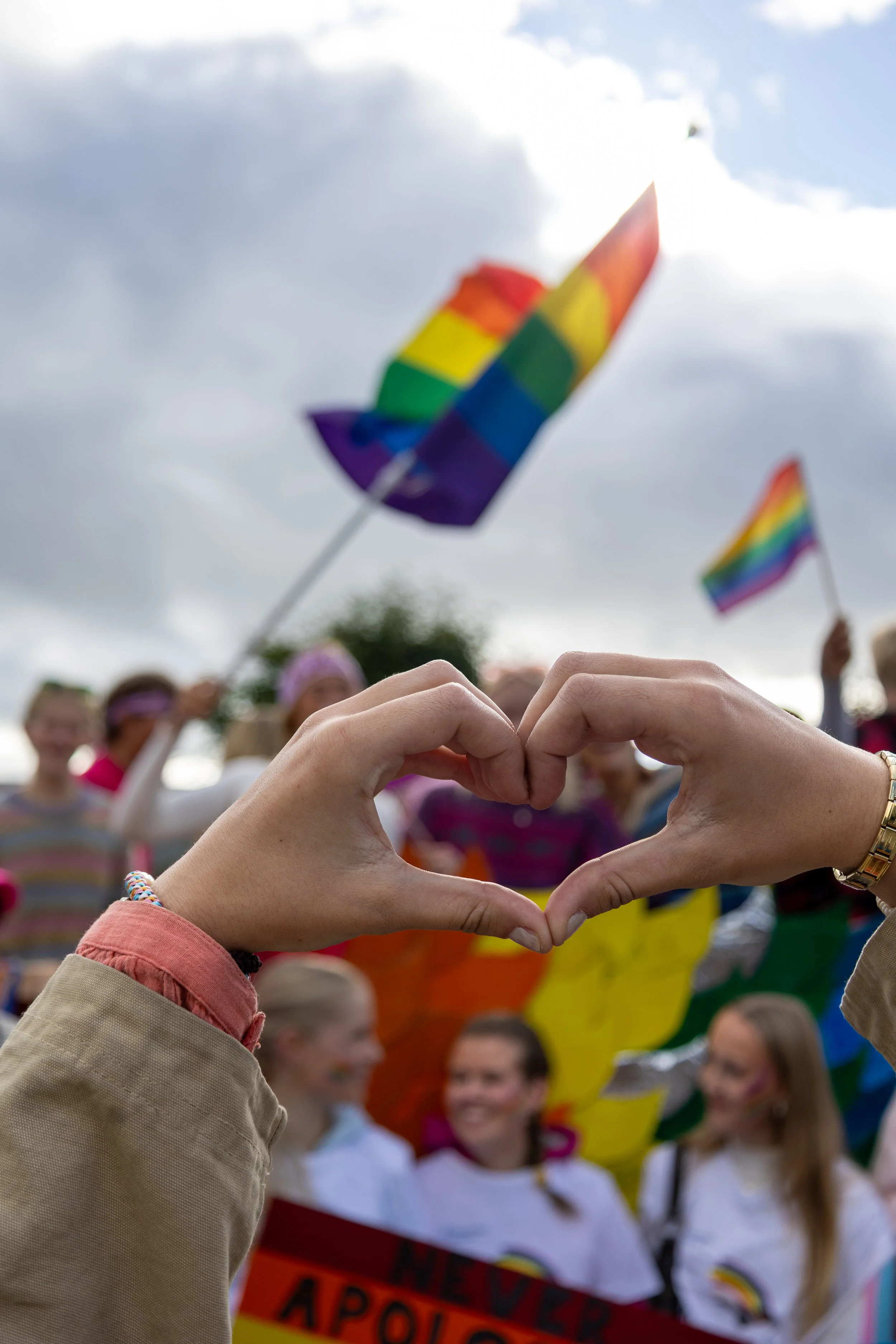 To hender danner et hjerteformet symbol foran en gruppe mennesker på en fargeglad parade med regnbueflagg i bakgrunnen.