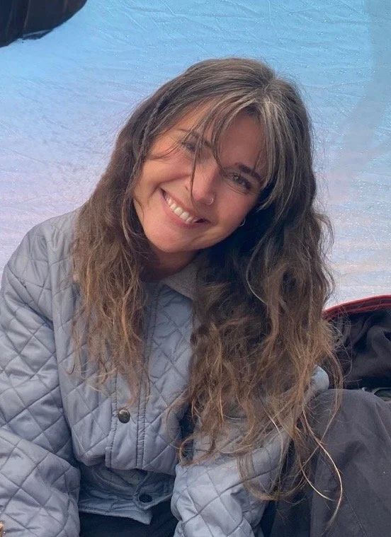 Kvinne med langt, bølgete brunt hår smiler og ser mot kamera, iført en lysegrå quiltet jakke. Bakgrunnen er en blå overflate.