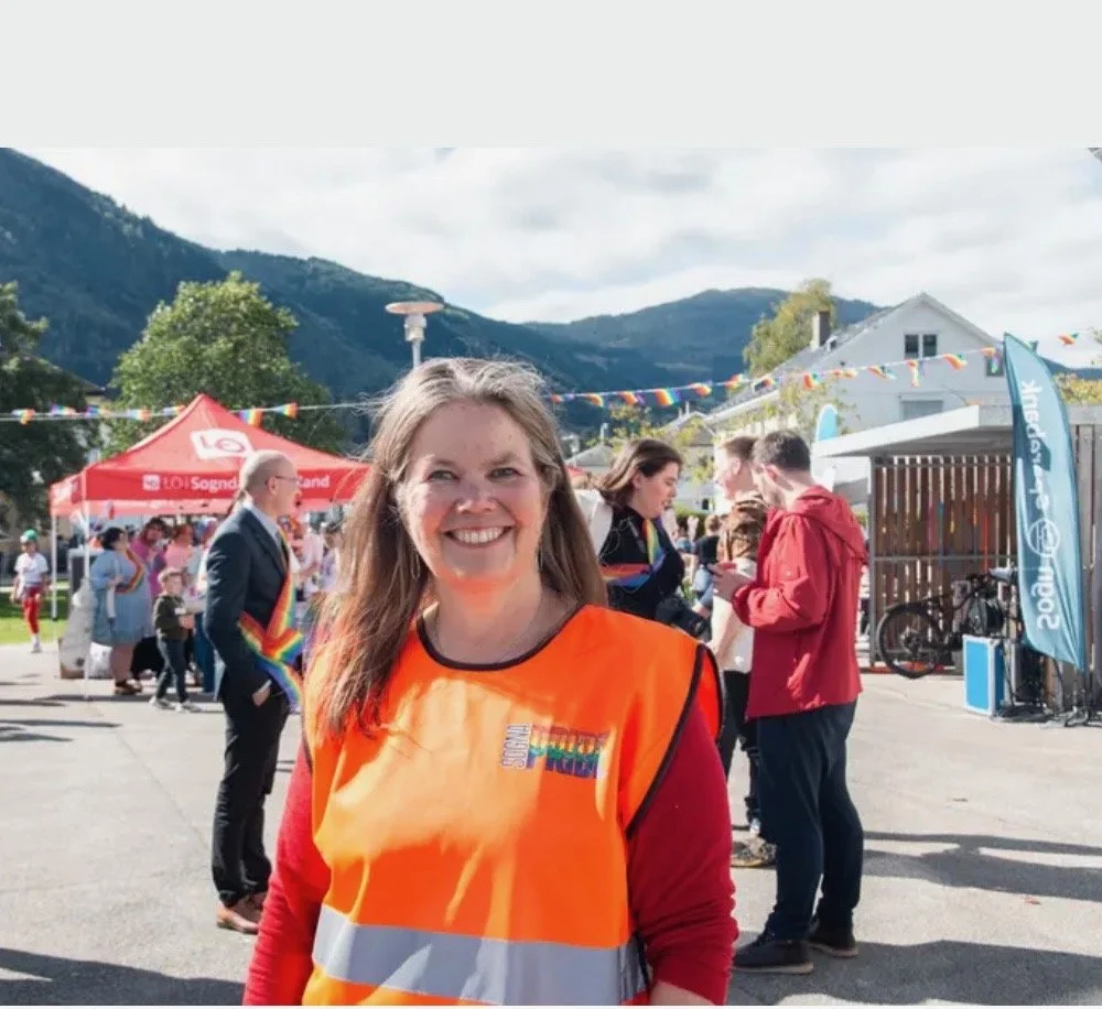 En smilende kvinne som deltar i en utendørs arrangement, iført oransje vest med tekst, omgitt av mennesker, boder, flagg og fjell i bakgrunnen.