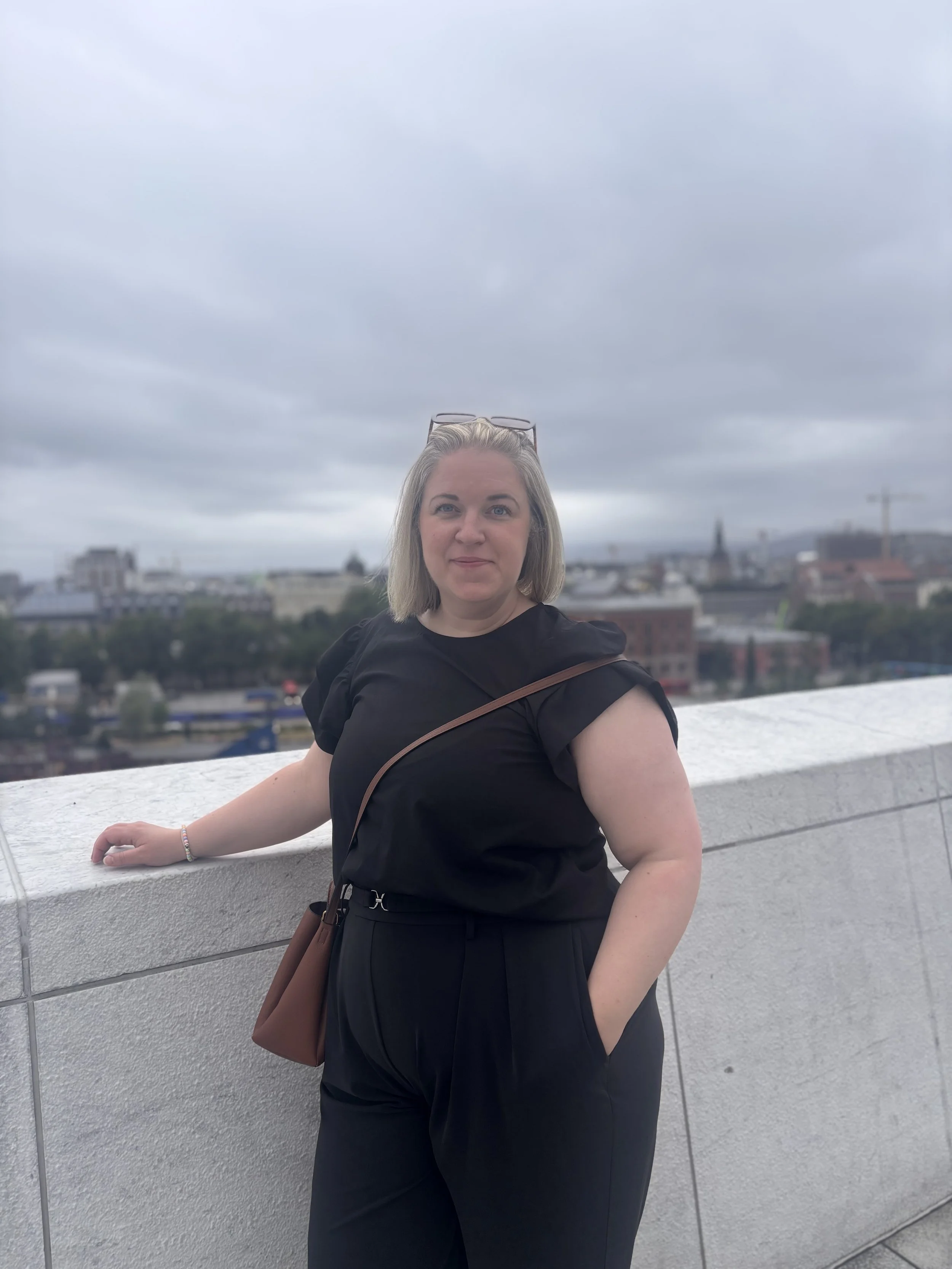 Ung kvinne med blond hår, svart topp og sorte bukser, står på en takterrasse med utsikt over en by, overskyet vær.