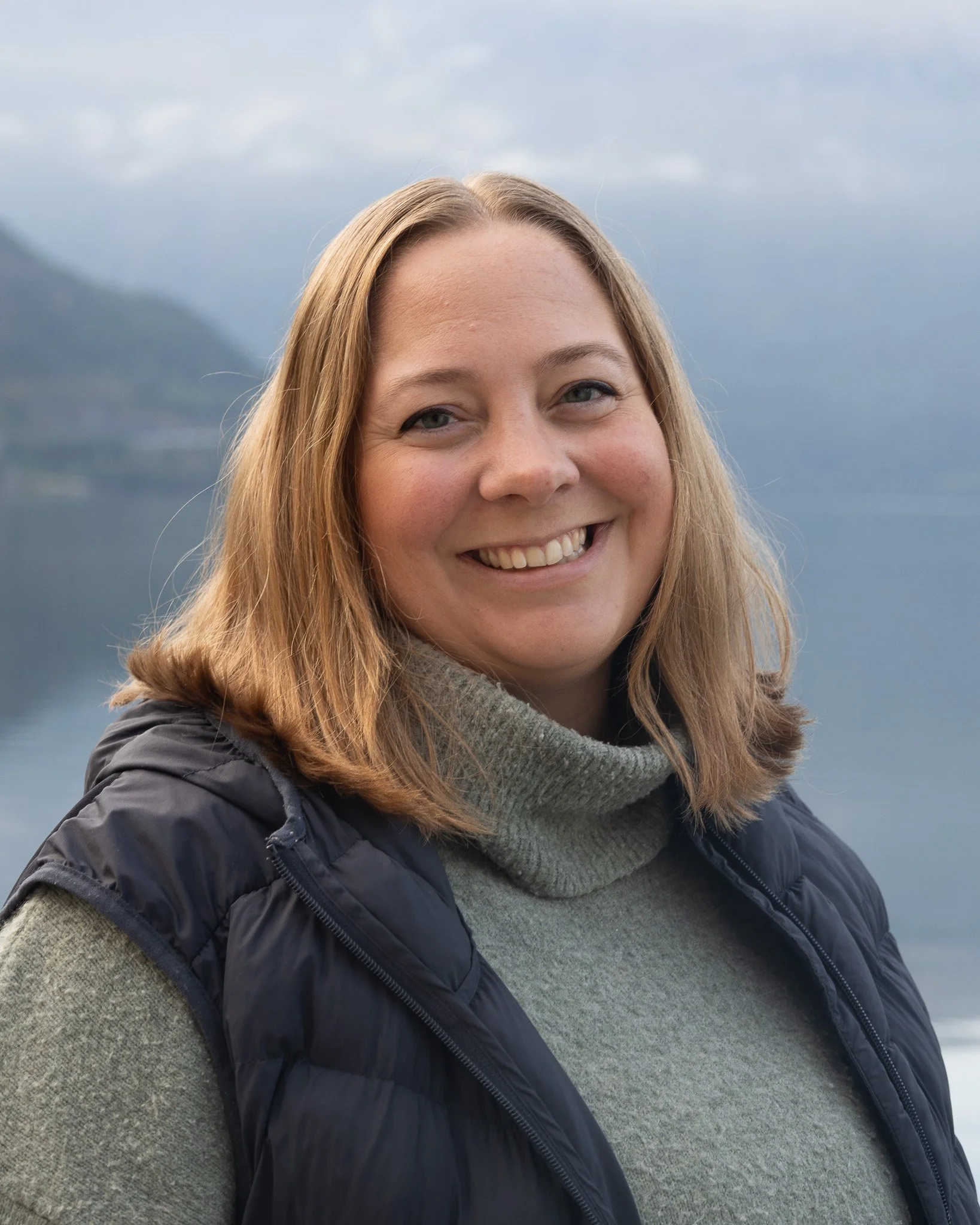Portrett av en kvinne med smil, ved vann, med fjell i bakgrunnen, iført grå genser og svart vest.