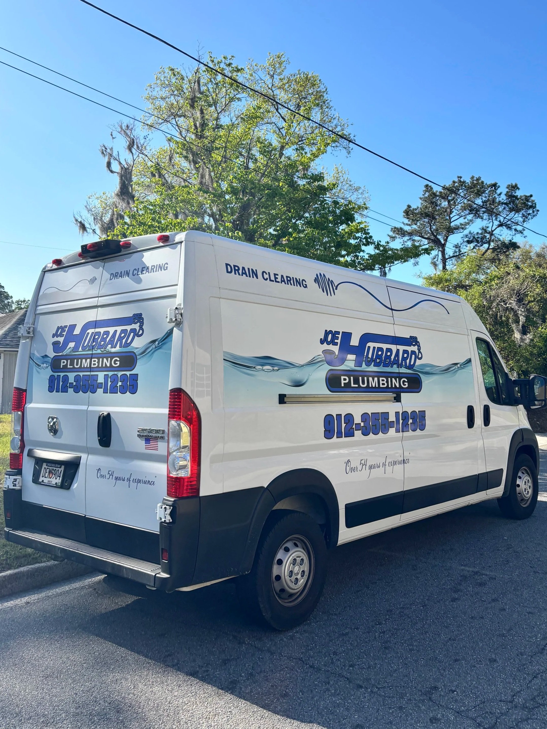 A white plumbing service van parked on the side of a street with trees and power lines overhead. The van has company branding that reads 'Joe Hubbard Plumbing' and a phone number, along with the phrase 'Over 54 years of experience.'