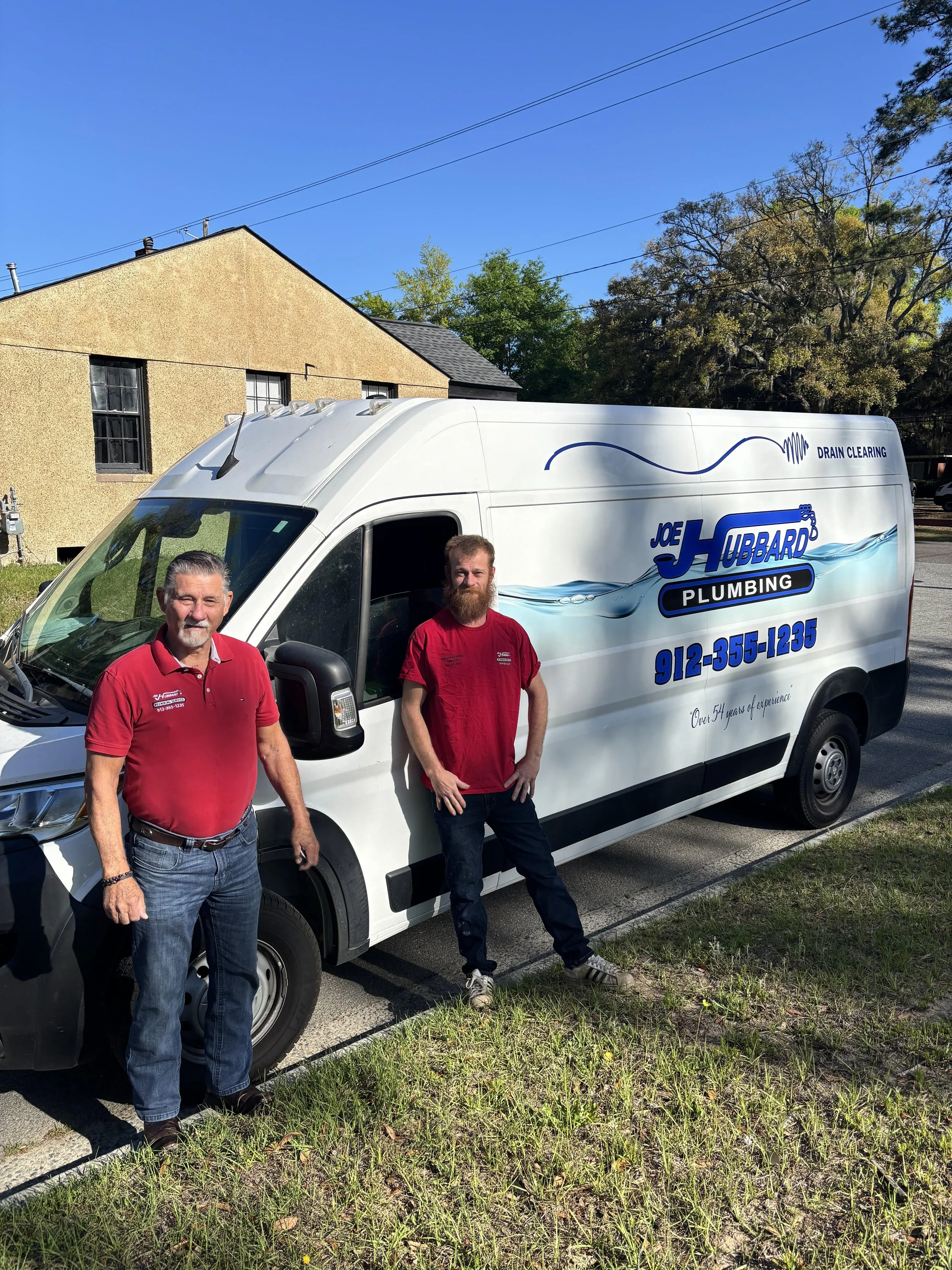 Two men standing in front of a white plumbing service van on a sunny day, with a beige building and trees in the background.