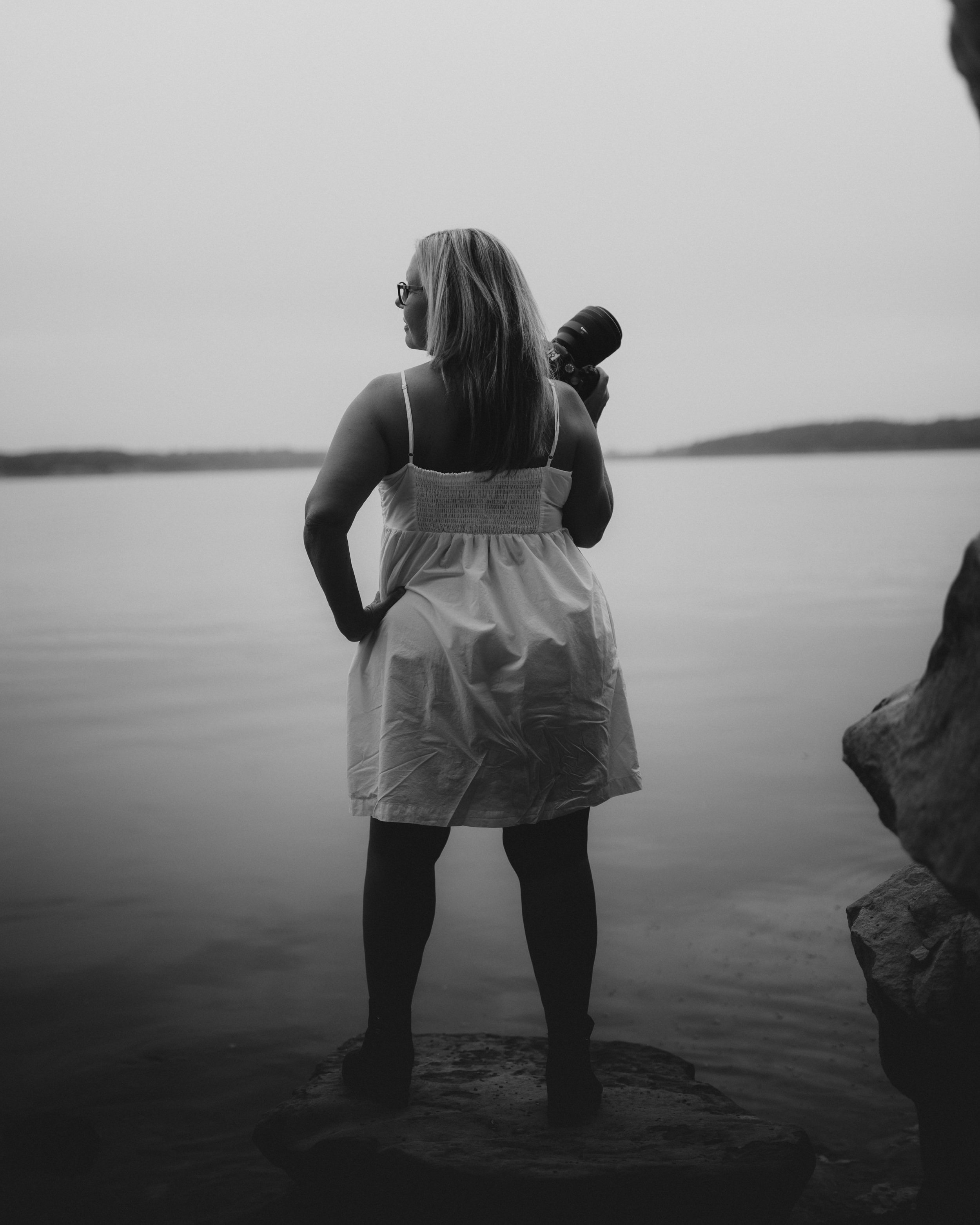 A woman with glasses in a white dress holding a camera, standing on a beach near the water with a distant landmass visible in the background, captured in black and white.