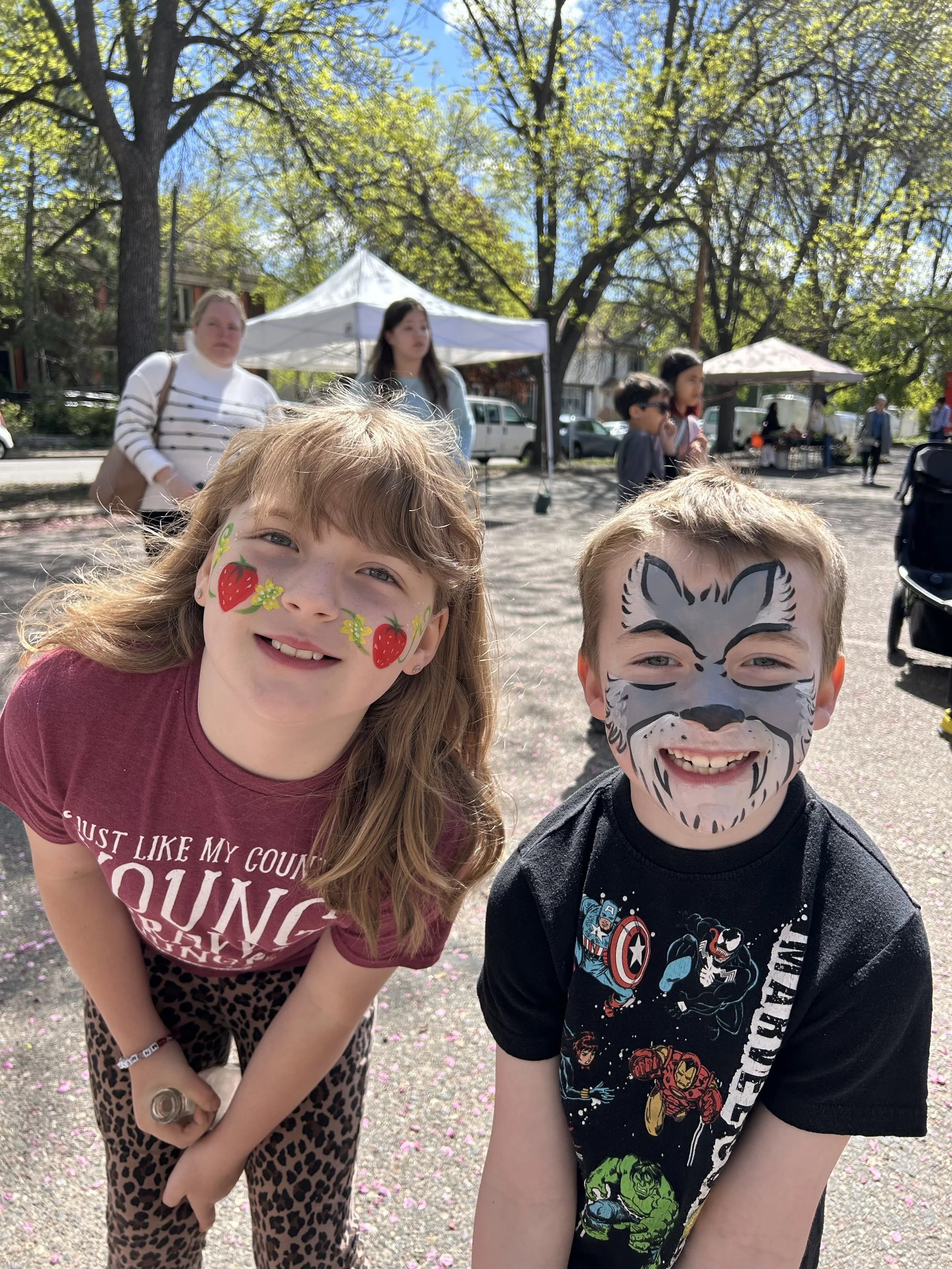 Two children with face paint, smiling, standing outdoors on a sunny day, with adults and trees in the background.