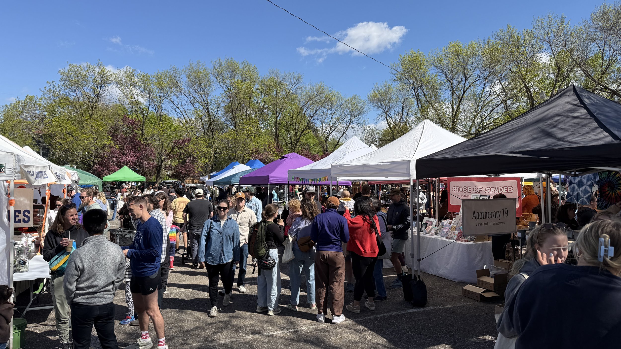Crowd of people at an outdoor market with tents and trees in the background on a sunny day.