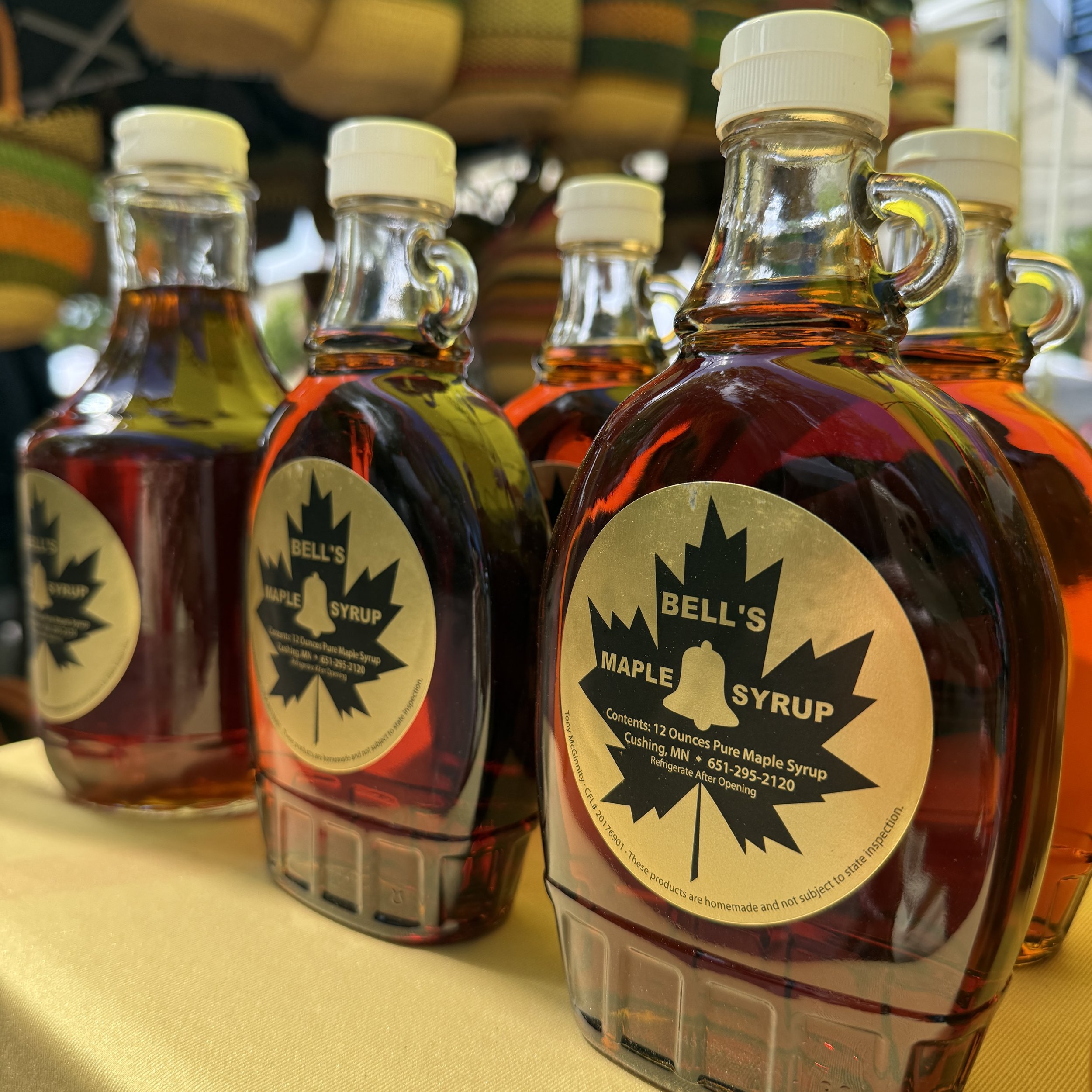 Several glass jars filled with maple syrup arranged on a yellow table, with a wooden display of more jars in the background.