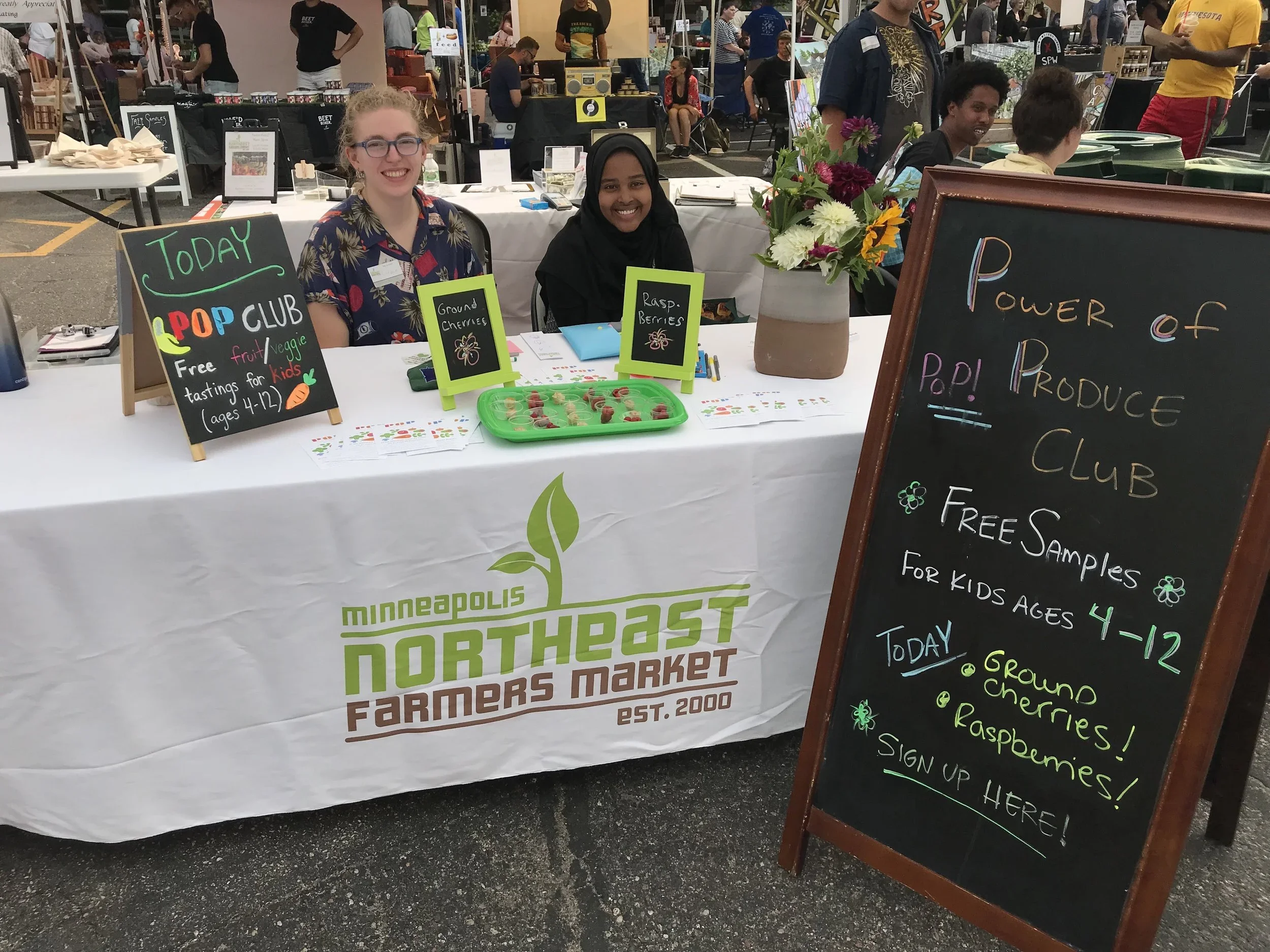 Two women sit behind a table displaying small samples of fruit, with signs indicating they are selling ground cherries, raspberries, and cherries at a farmers market. A floral arrangement is on the table, and there are other market booths and people visible in the background.