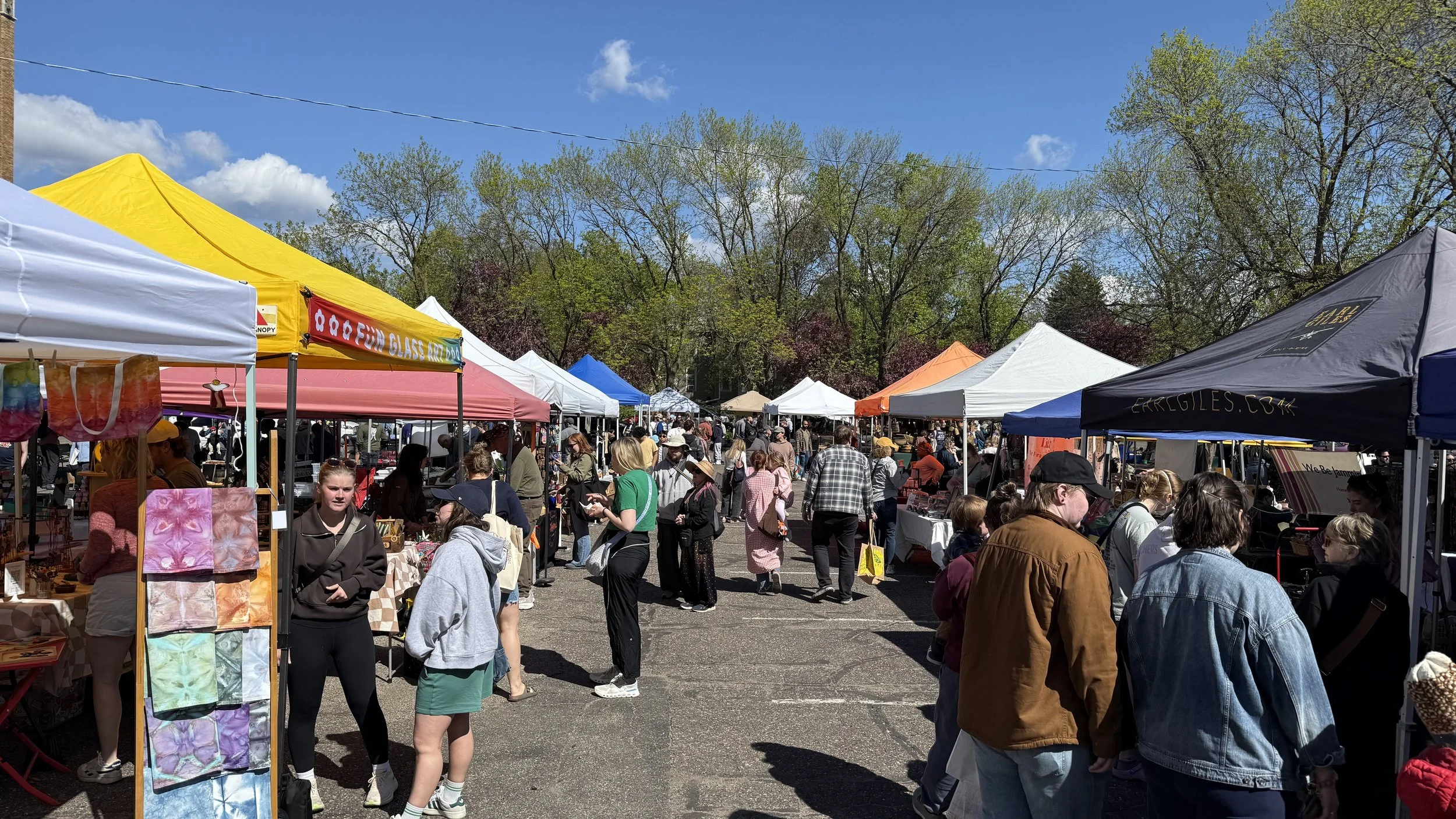 A crowded outdoor market with colorful tents and vendors selling various items, with many people browsing and shopping on a sunny day with blue skies and trees in the background.