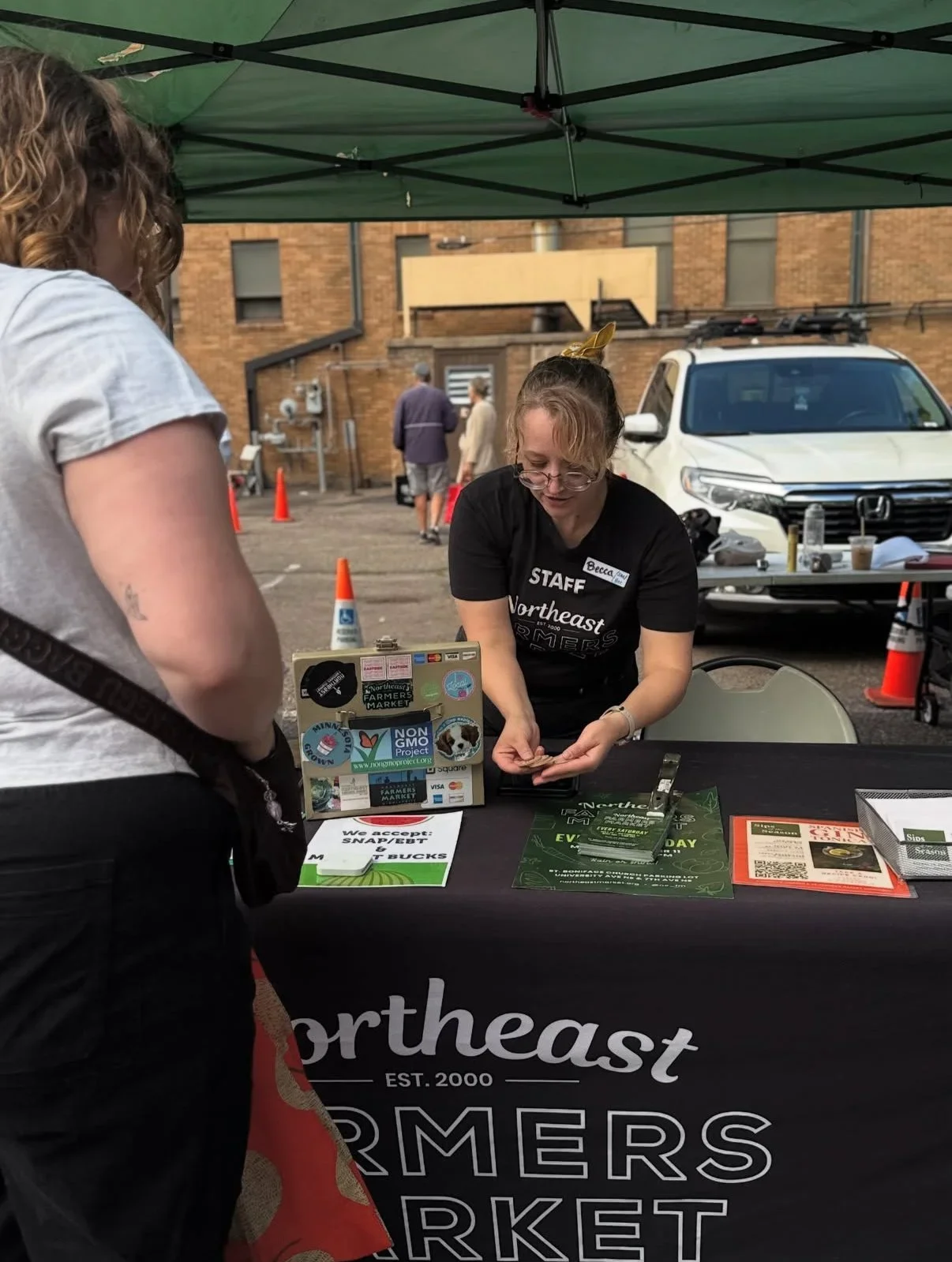 A woman with glasses and a black t-shirt labeled "STAFF Northeast Farmers Market" is interacting with a customer at an outdoor market booth. The booth has a black tablecloth with the market's name and promotional materials, including stickers, flyers, and a small display stand. The setting is outdoors with a green canopy overhead, brick buildings, and parked cars in the background.