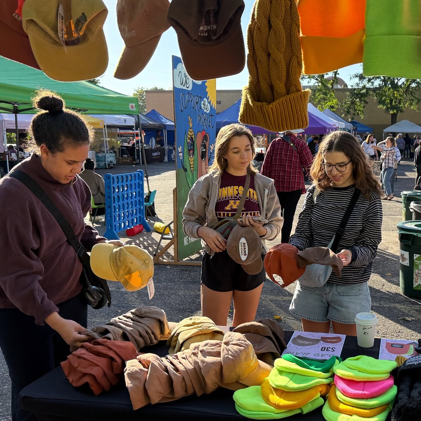 People shopping at an outdoor market with hats, scarves, and beanies on display.