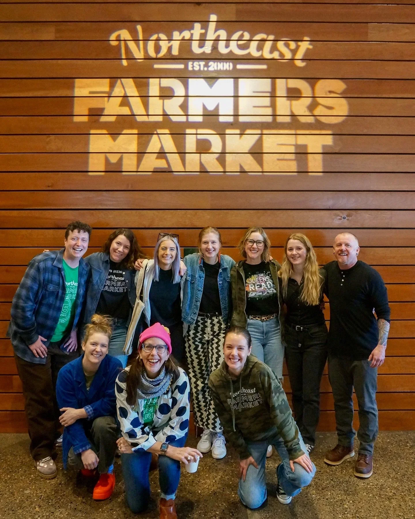 A group of people standing and kneeling in front of a wooden wall with a sign that reads 'Northeast Farmers Market.' The group includes men and women of various ages, some smiling and wearing casual clothing, with some wearing glasses and one person wearing a pink hat. The setting appears to be indoors.