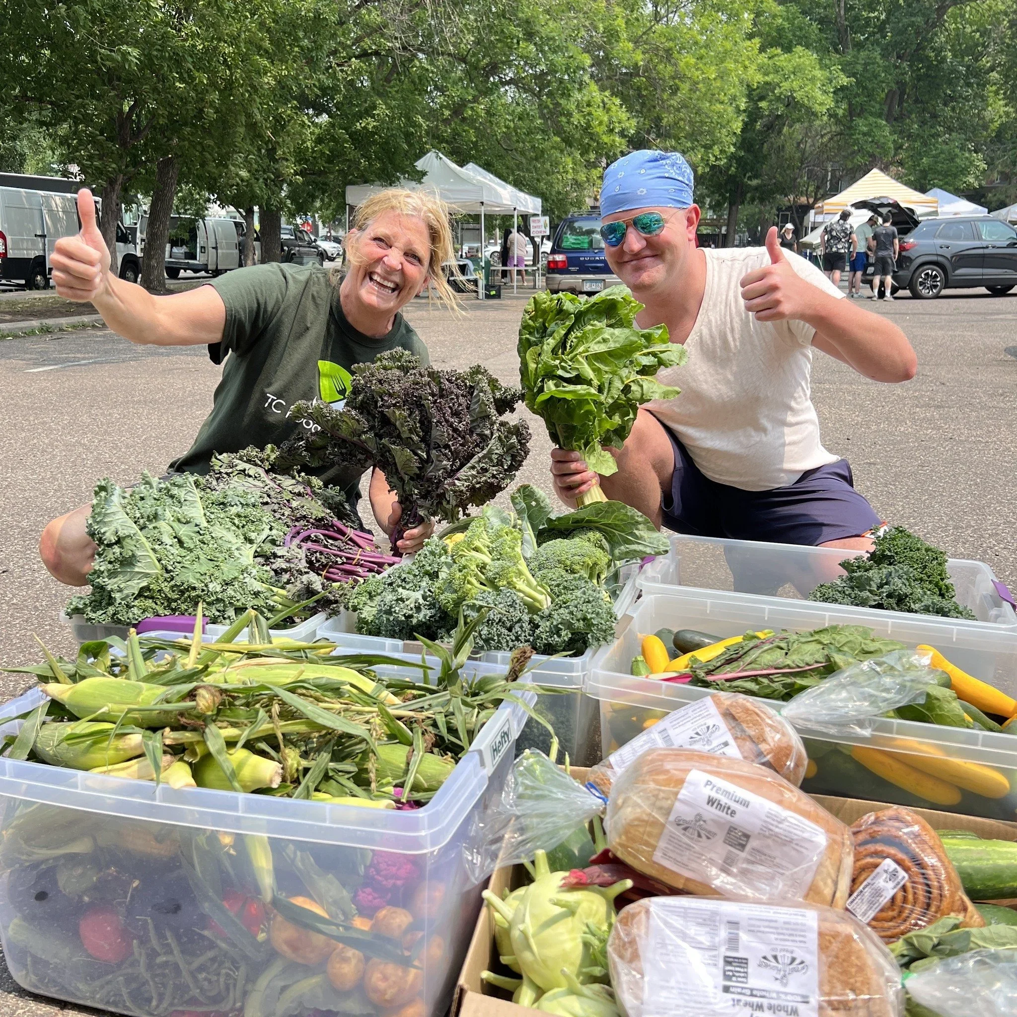 Two smiling people holding fresh vegetables at an outdoor market, with trees, tents, and parked cars in the background.