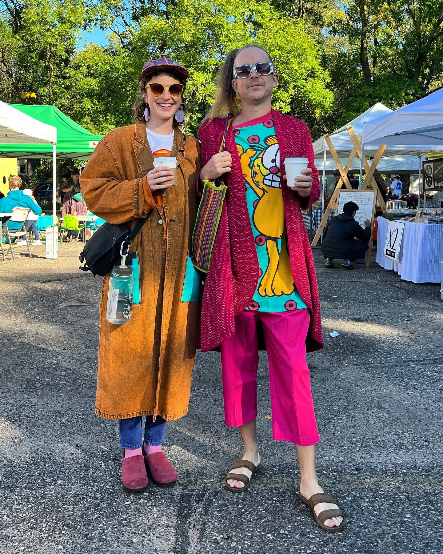 Two people dressed in colorful, eclectic outfits standing outdoors with green trees and market stalls in the background, holding coffee cups and smiling.