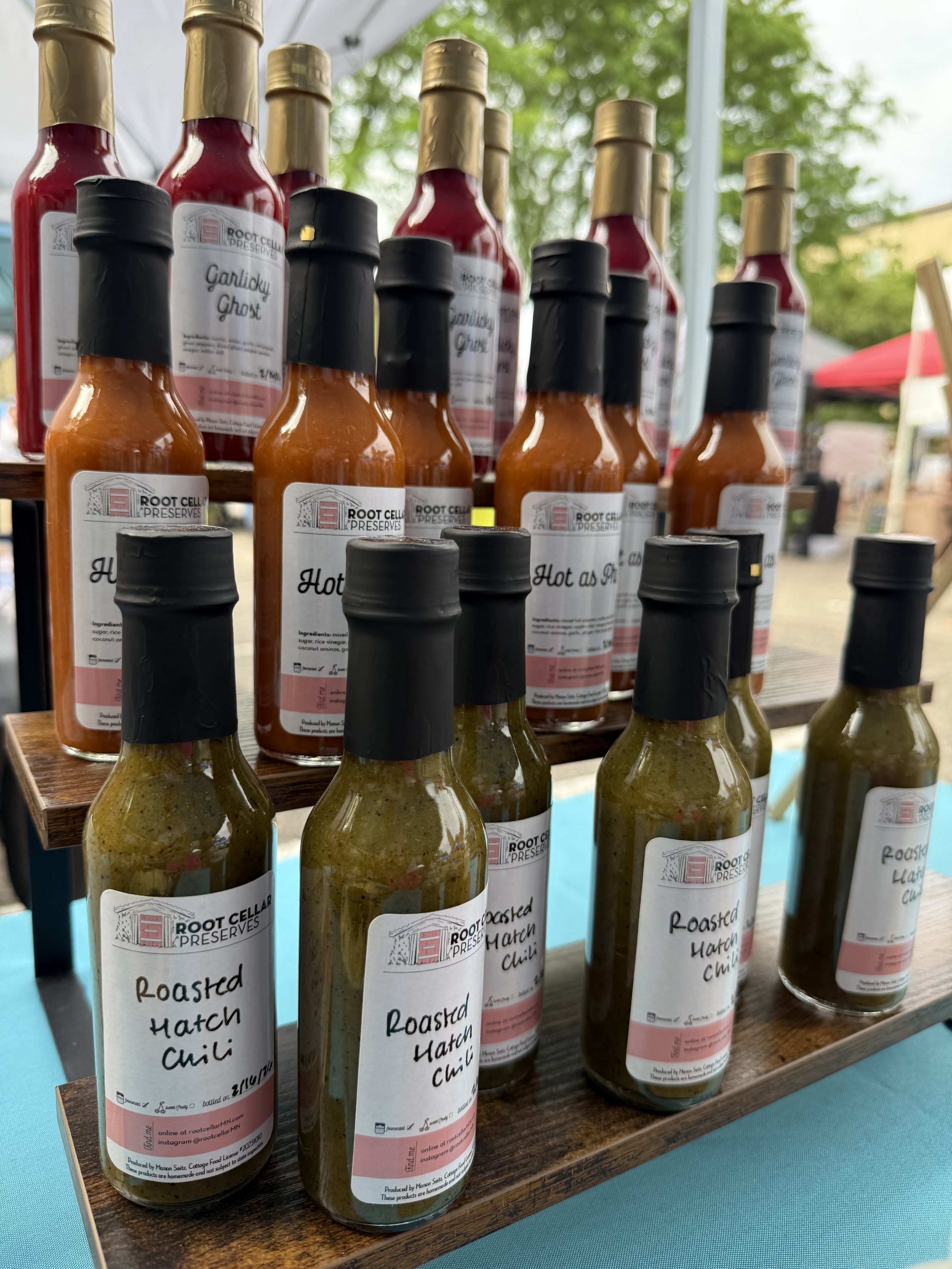 Bottles of hot sauce labeled 'Roasted Hatch Chili' displayed on a wooden stand at an outdoor market.
